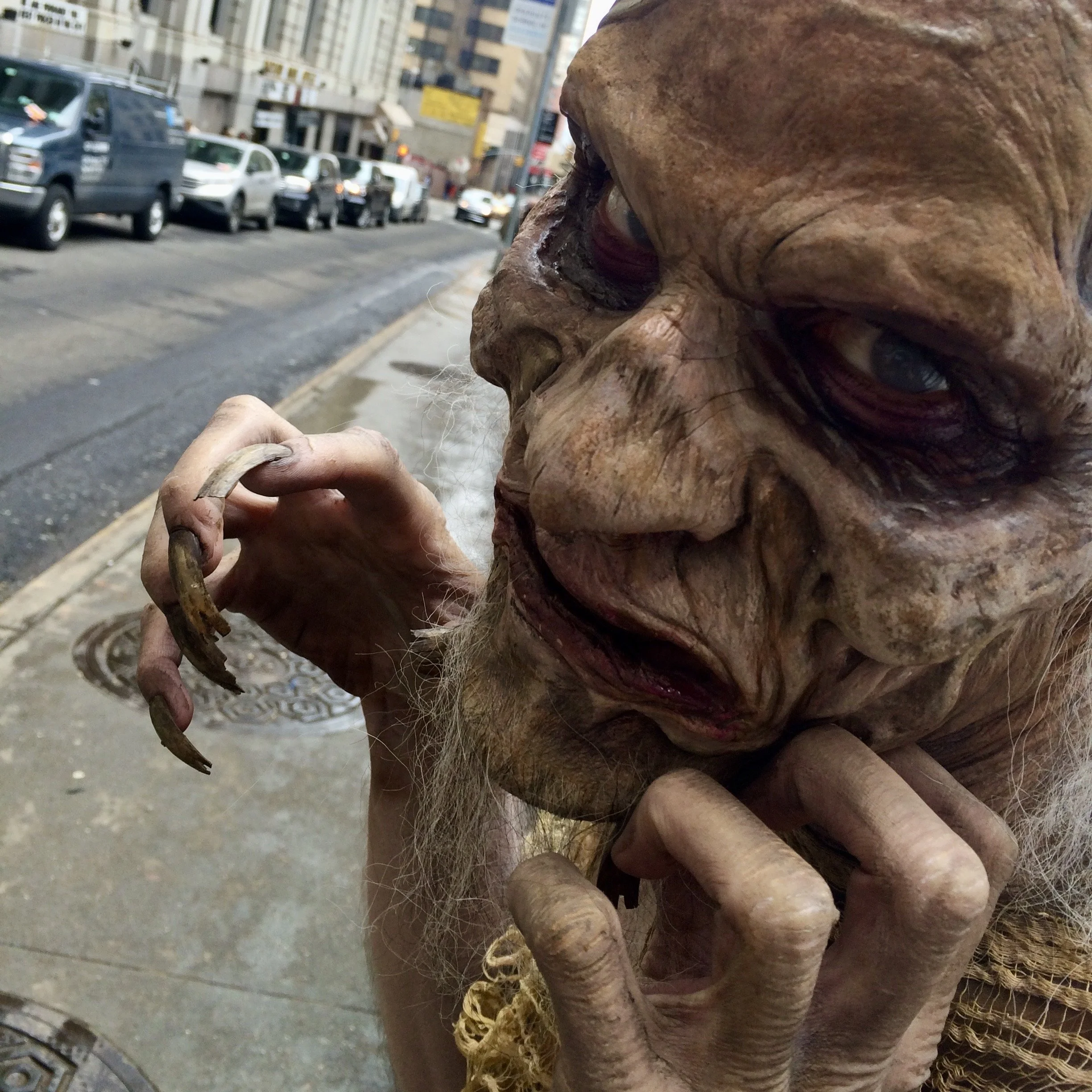 Close-up of a creature with wrinkled, mottled skin, long claws, and eerie eyes, standing on a city street with cars and buildings in the background.