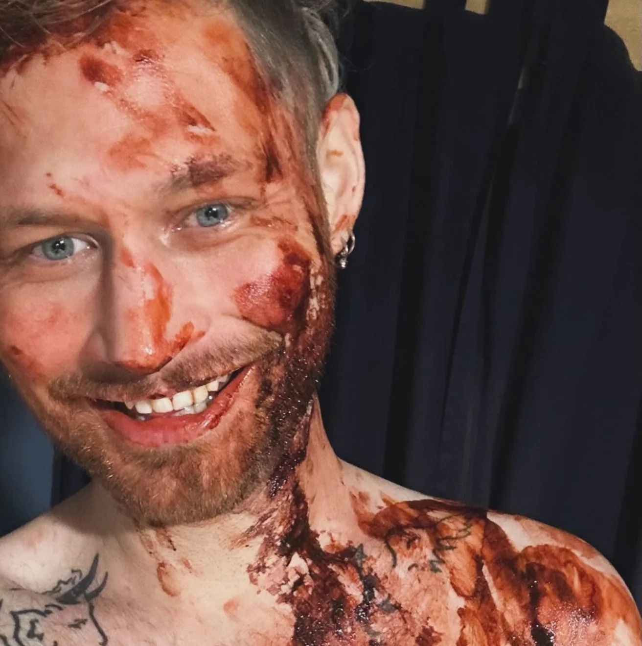 A man with blue eyes and short hair, covered in fake blood and wounds, smiling and showing his teeth.