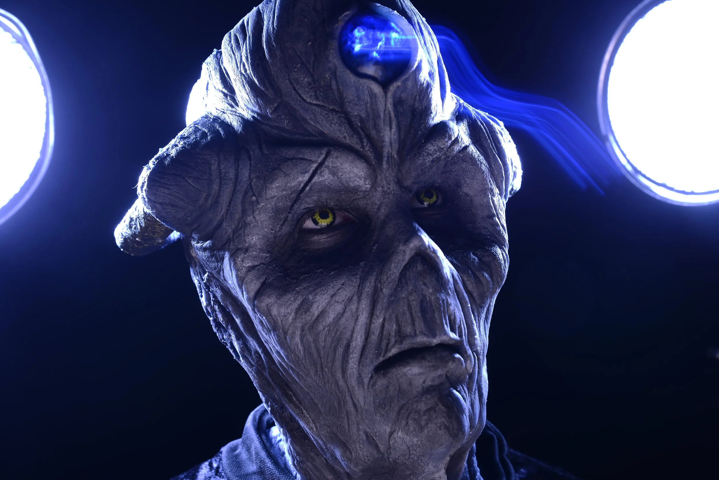 Close-up of an alien creature with gray, wrinkled skin, yellow eyes, and two curled horns, illuminated by bright white lights in the background.