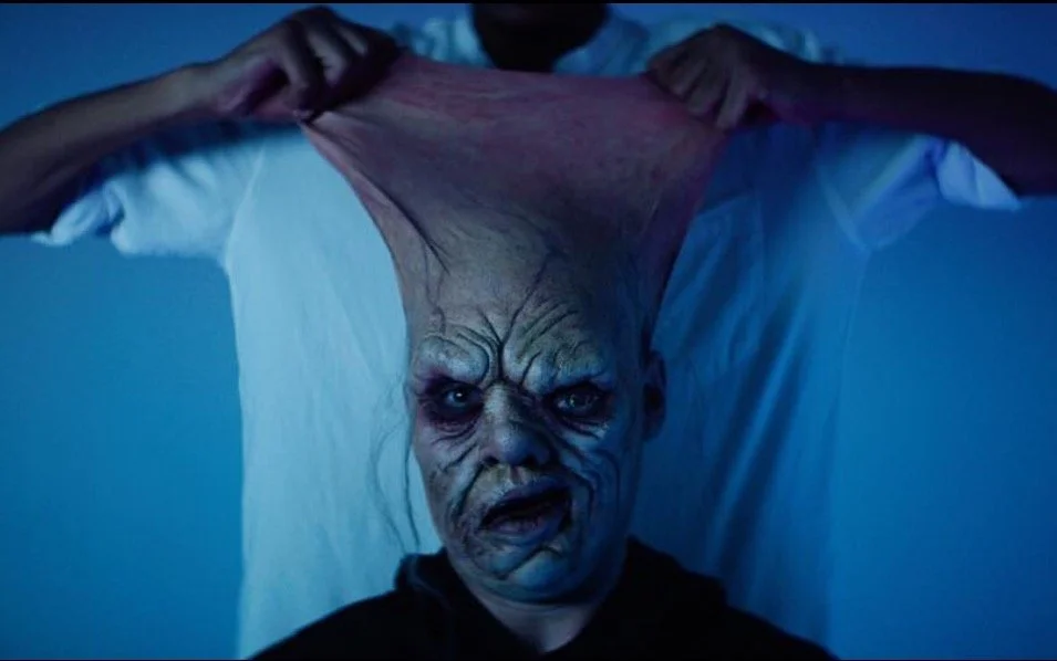 A person with a distorted face pulling a stretchy skin-like mask over their head in a creepy setting with blue lighting.