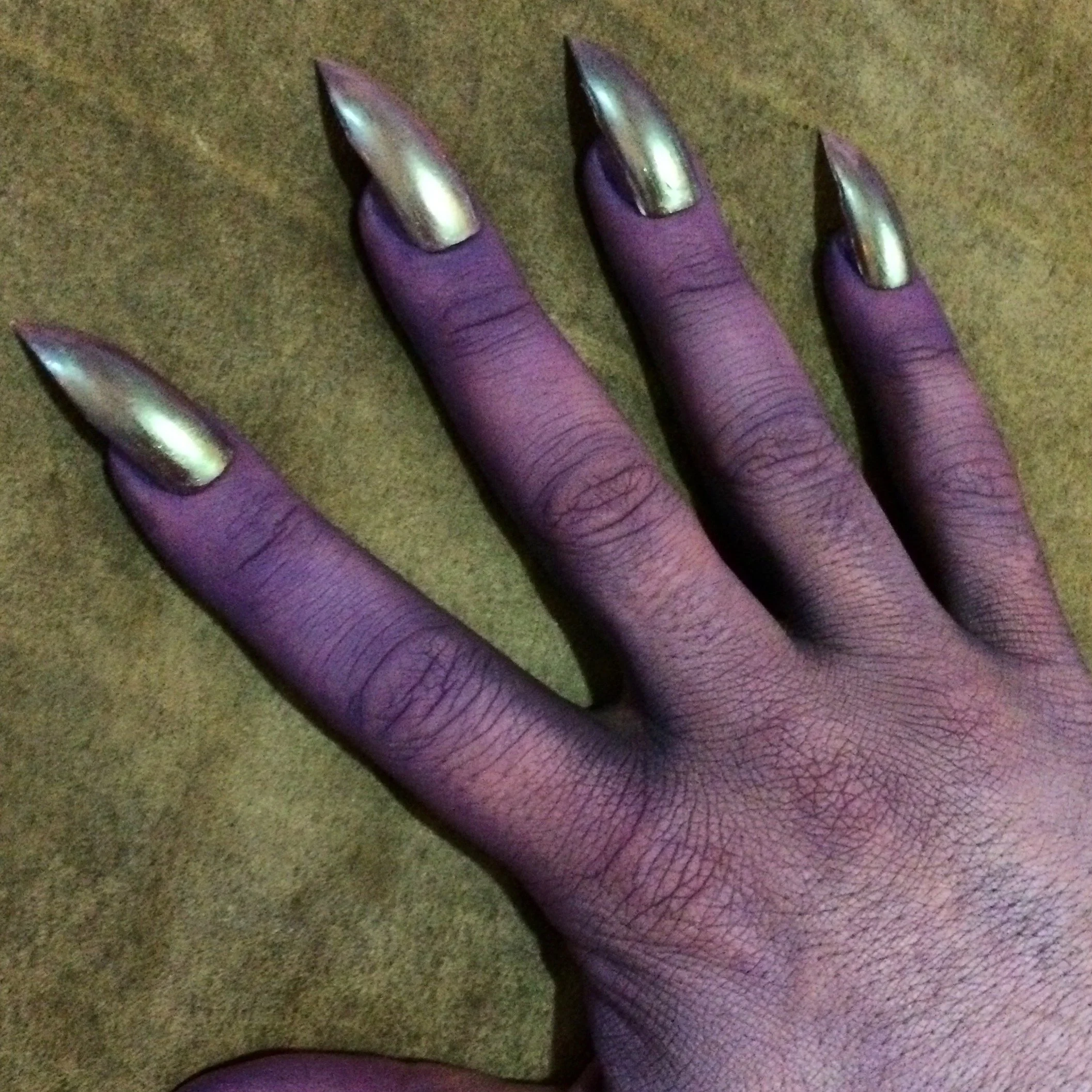 A hand with dark skin tone and long, pointed metallic nails, resting on a brown textured surface.