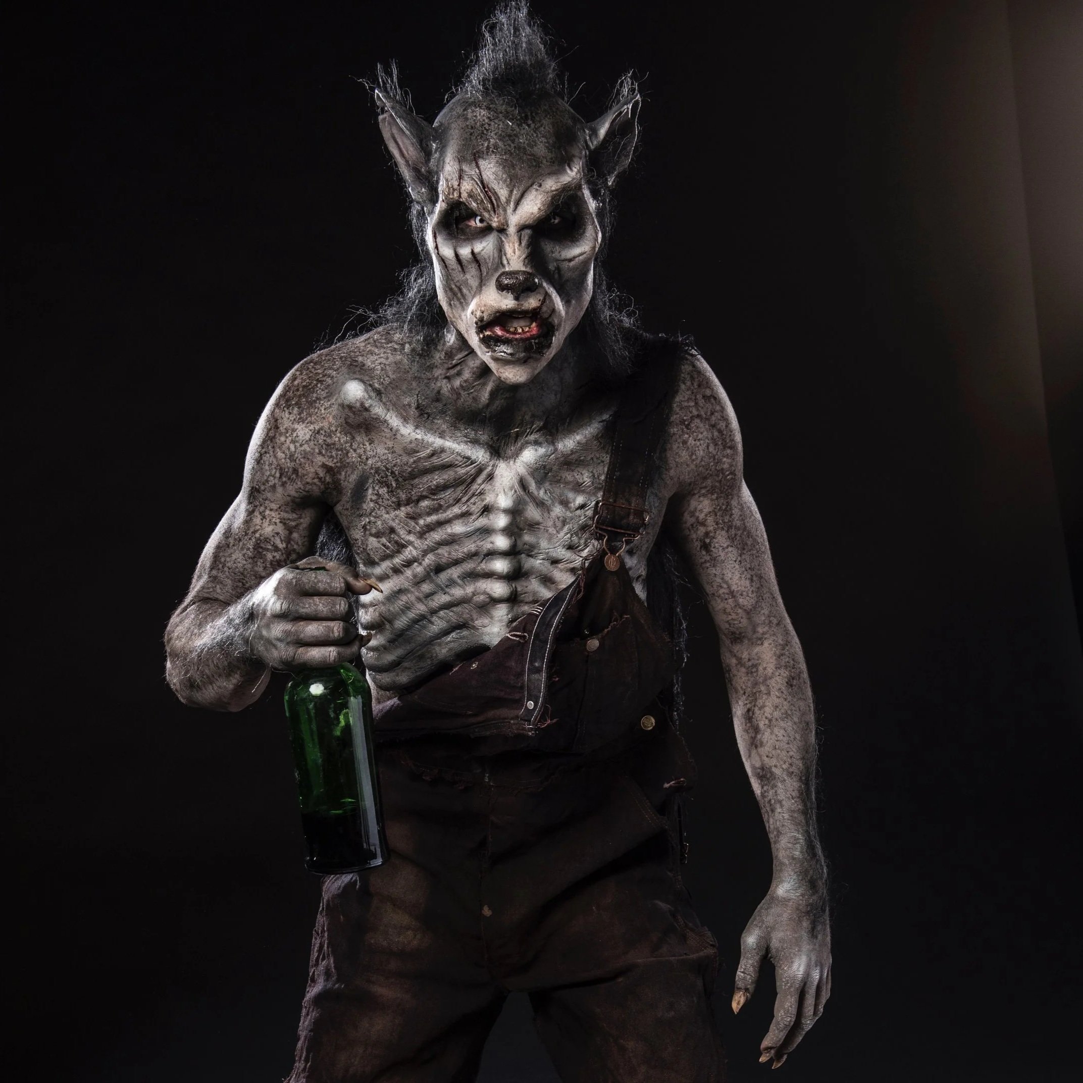 A person dressed in a werewolf costume with gray fur, animal-like face makeup, and claws, holding a green bottle, against a black background.