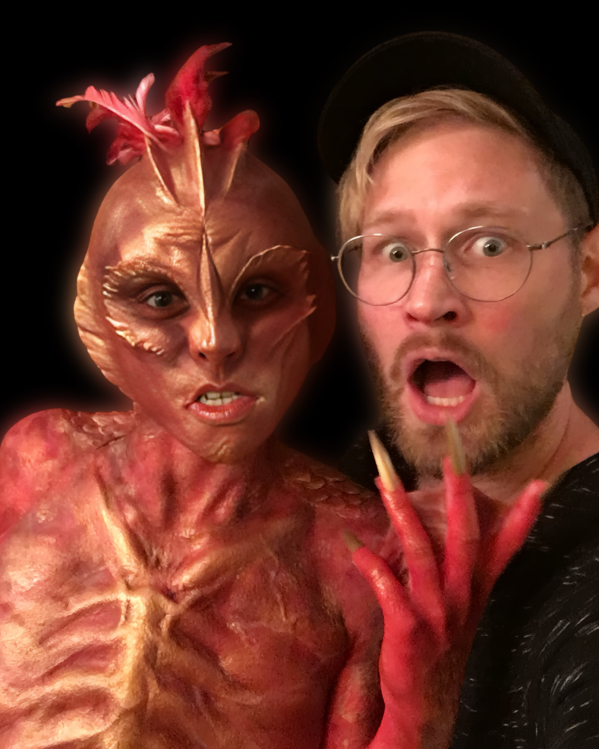 A person in a detailed crab costume with an armored body, crab claws on their head, and textured body, taking a selfie with a man wearing glasses and a black hat, both with surprised expressions.