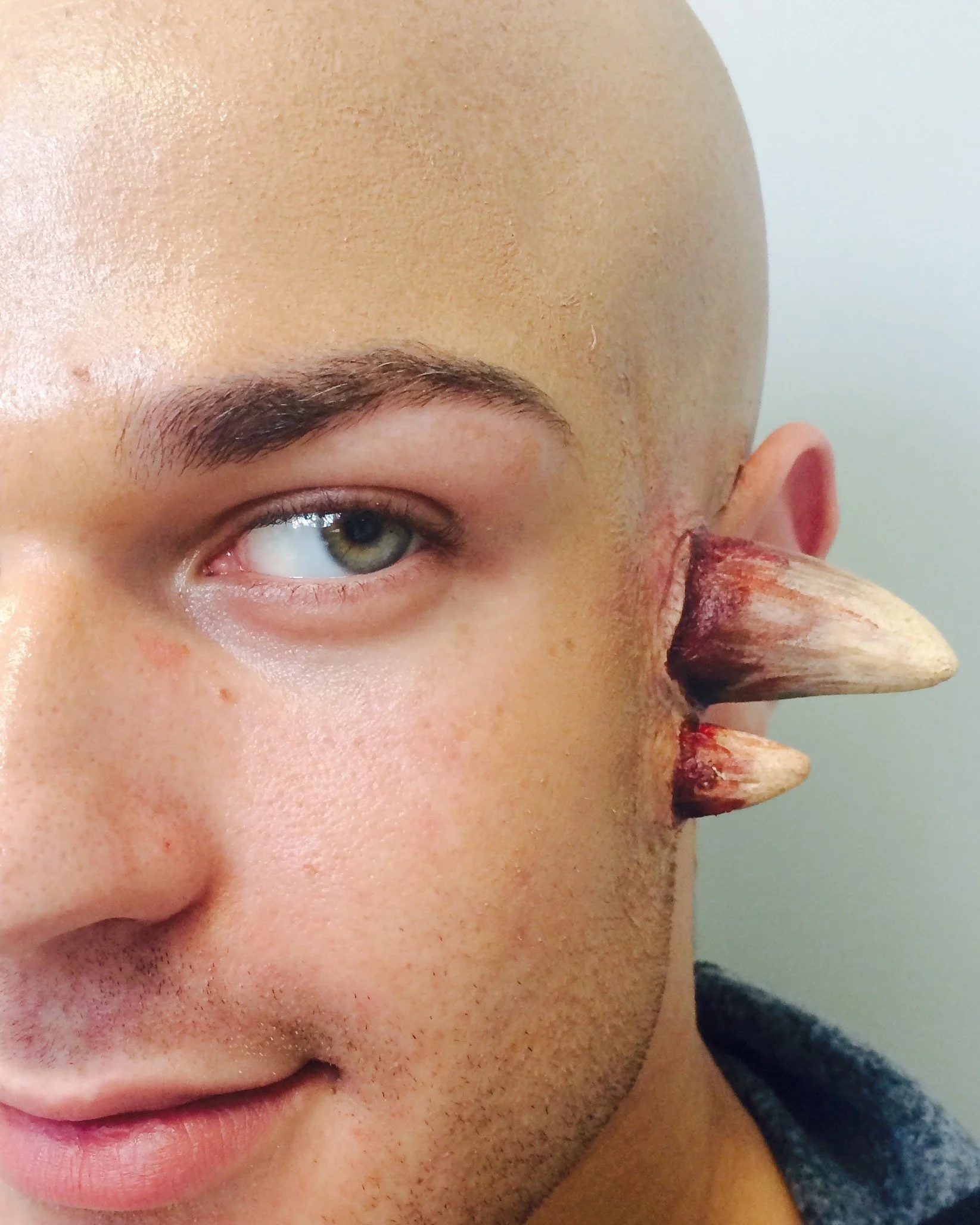 A person with fantasy makeup and costume, including fake animal-like teeth and a bald head, close-up of their face.