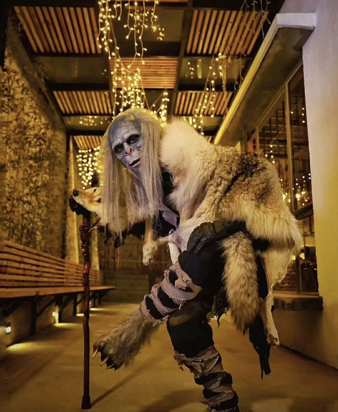 A person dressed as an orc or fantasy creature with long hair, face makeup, furry clothing, and rugged pants, holding a staff with a wolf head, in a decorated outdoor setting with string lights.