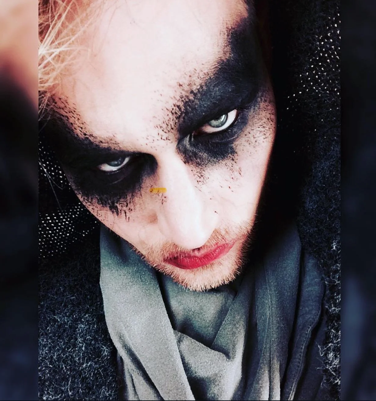 Close-up of a person with heavy black makeup around their eyes, pale skin, light-colored eyes, and wearing a dark hooded garment.