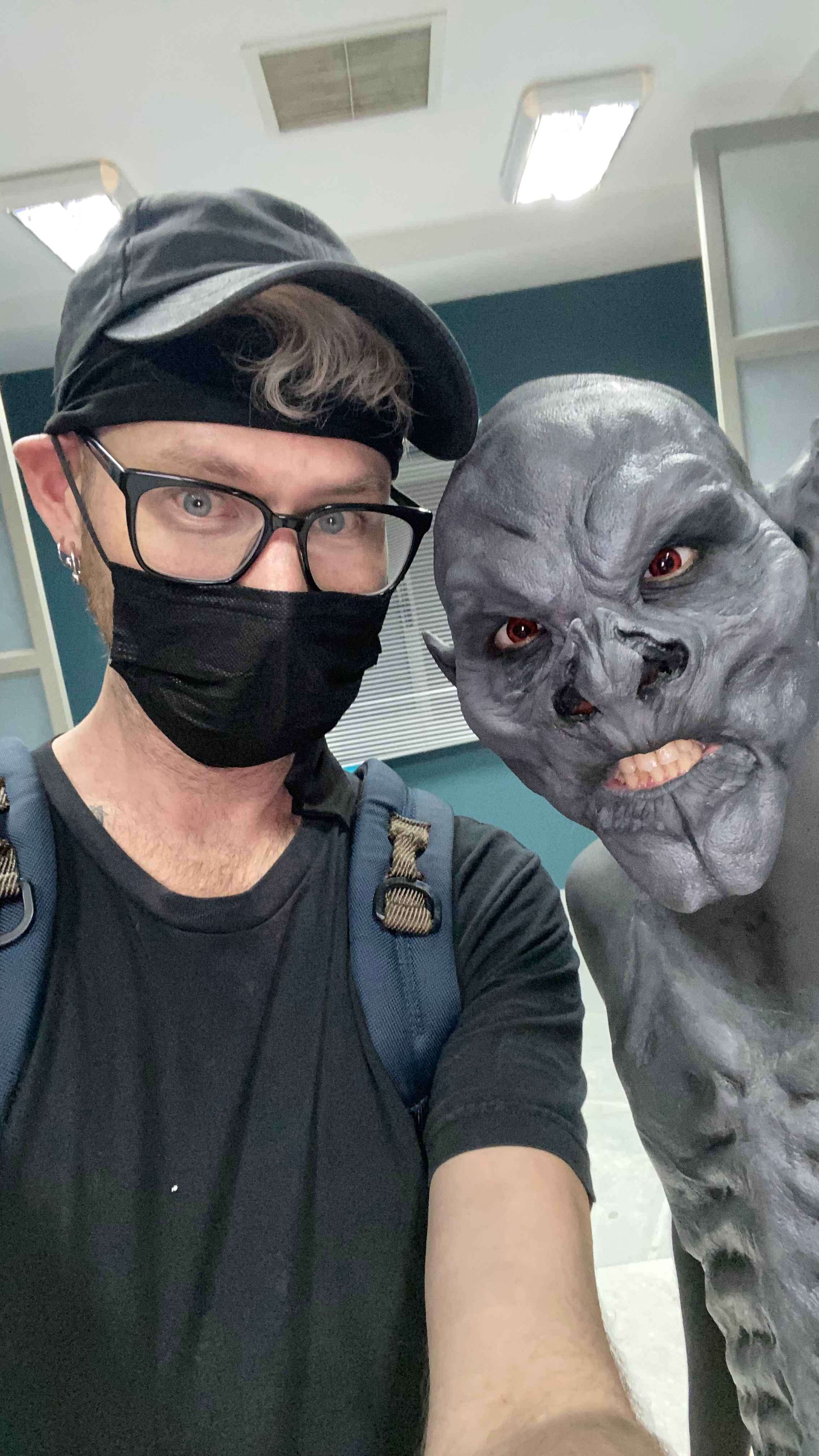 A person with glasses and a black face mask taking a selfie with a creepy gray creature that has orange eyes and a snarling expression.