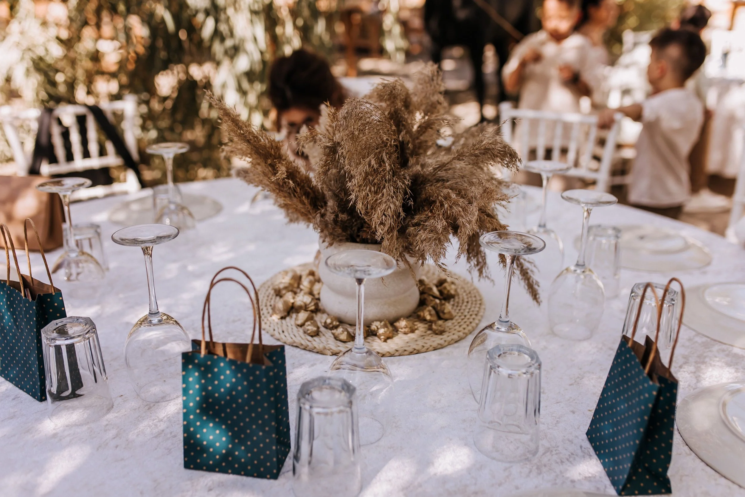 A table set for a celebration, decorated with a large floral centerpiece made of dried pampas grass in a white vase. The table has glass upside-down wine glasses, and small blue polka-dotted gift bags with brown paper handles around the edges. In the background, people are talking, and the setting appears to be outdoors with natural light.