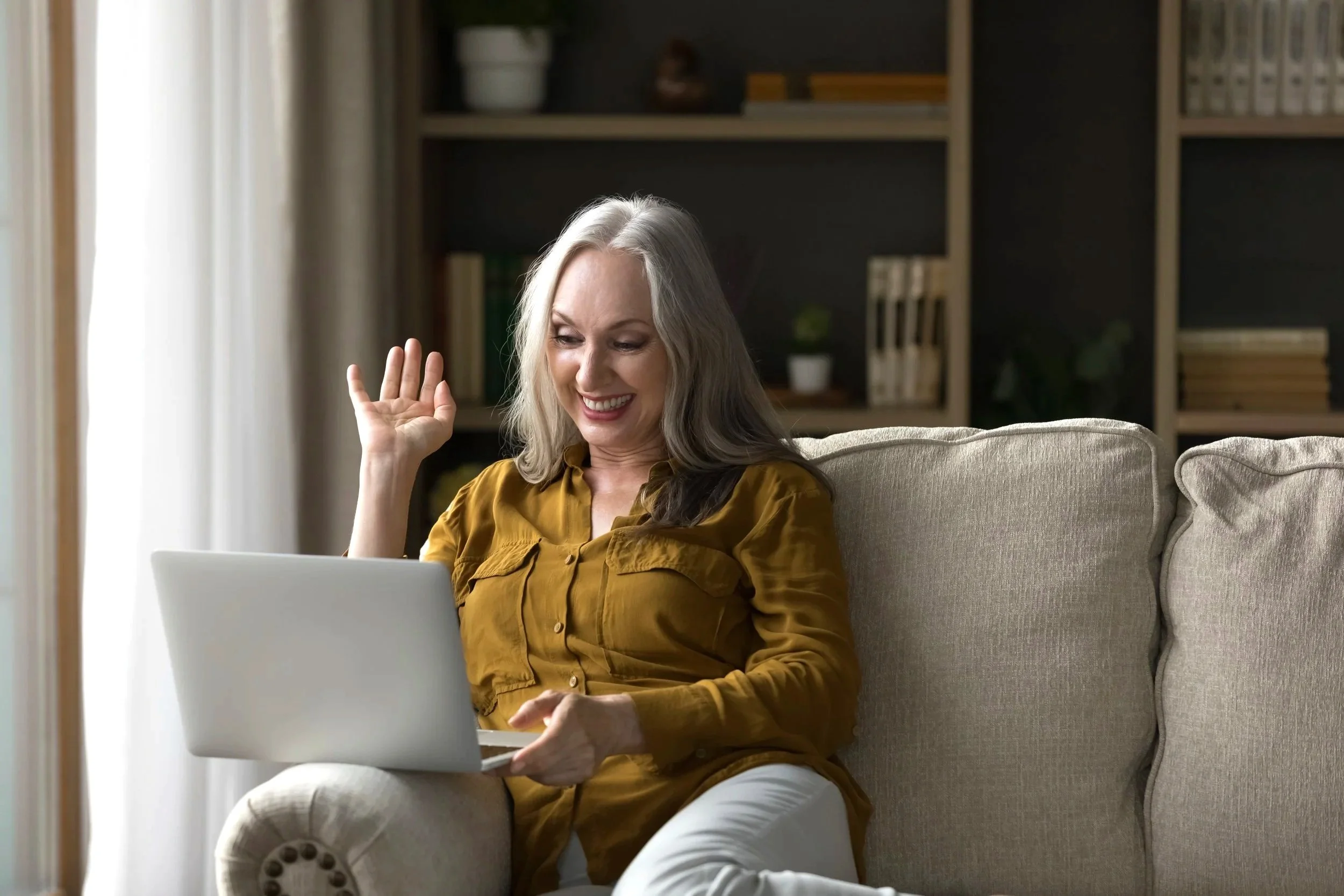 A smiling woman with gray hair sitting on a beige sofa, holding a laptop, and waving at the screen in a cozy living room with a bookshelf and curtains.