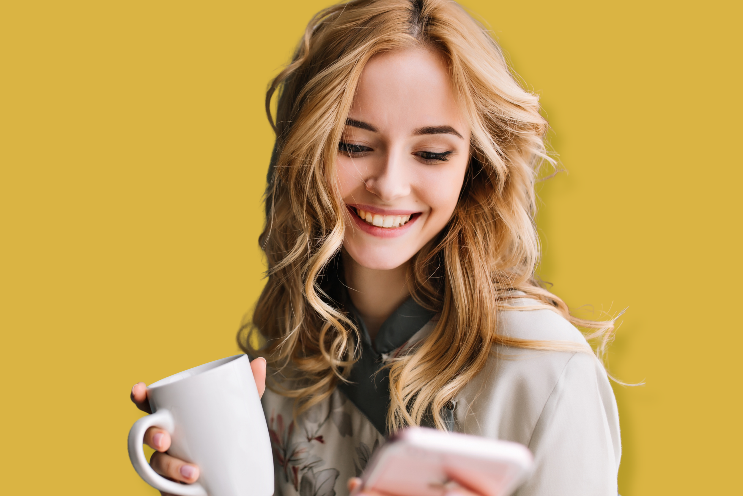 A young woman with blonde curly hair smiling while looking at her phone, holding a white mug, against a yellow background.