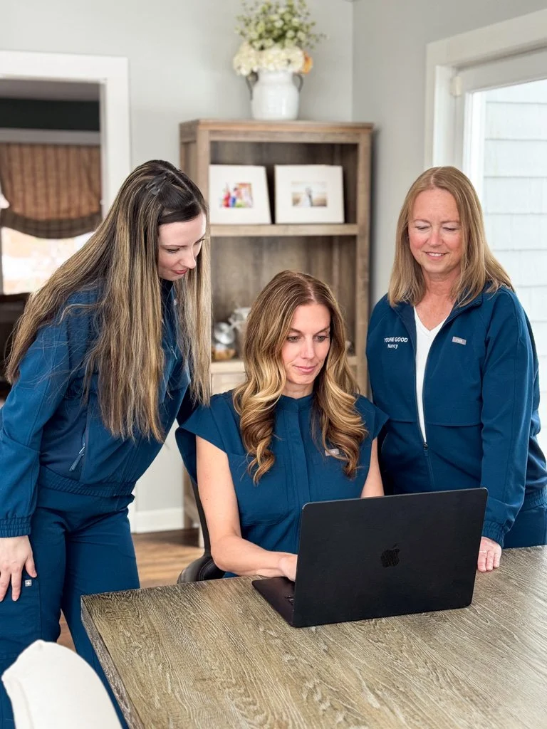 Three women in blue uniforms working together around a laptop in a bright room with a wooden table and shelves.