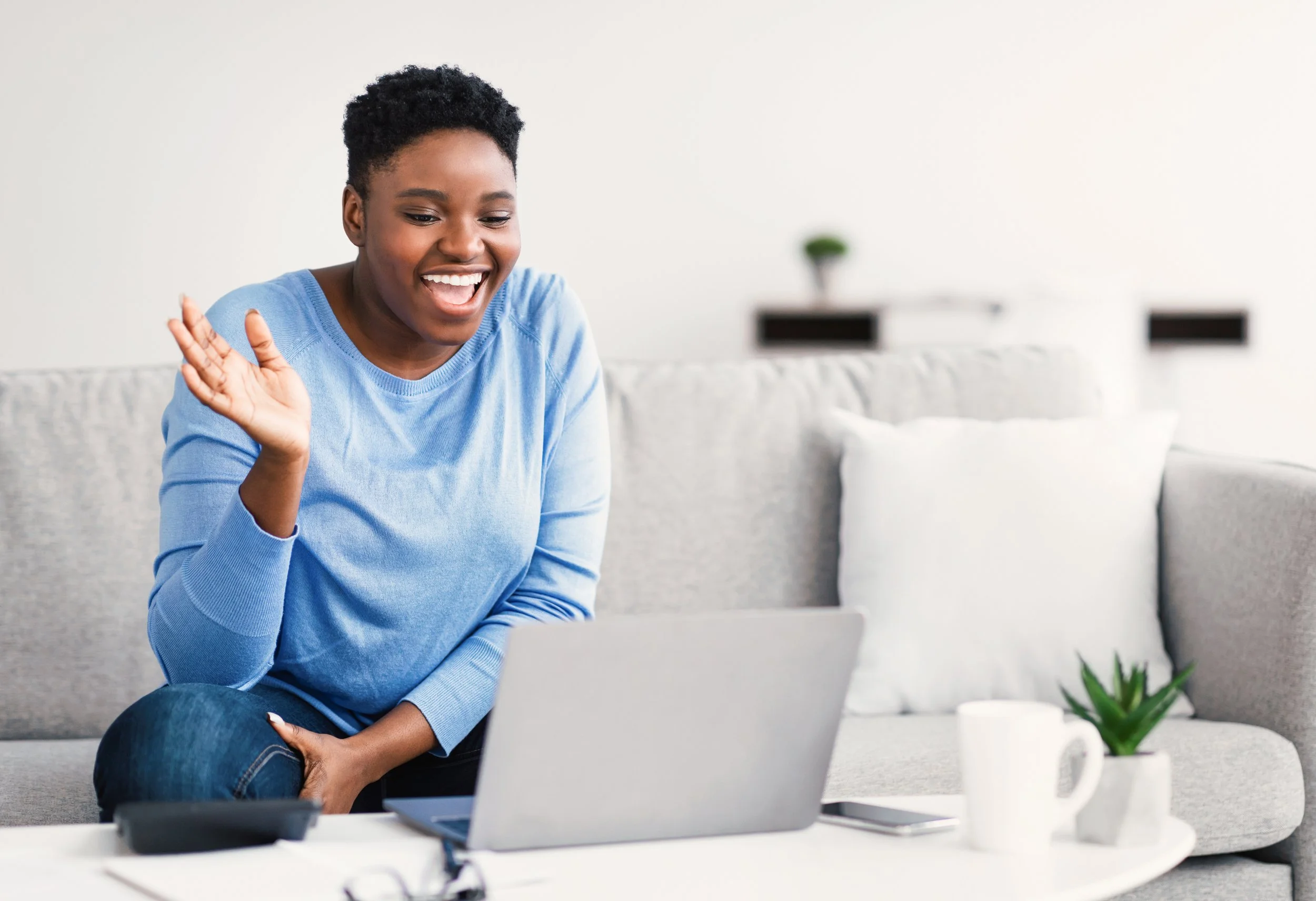 A woman with short black curly hair, wearing a light blue long-sleeve shirt, smiling and waving at her laptop while sitting on a beige sectional sofa in a bright living room with minimal decor.