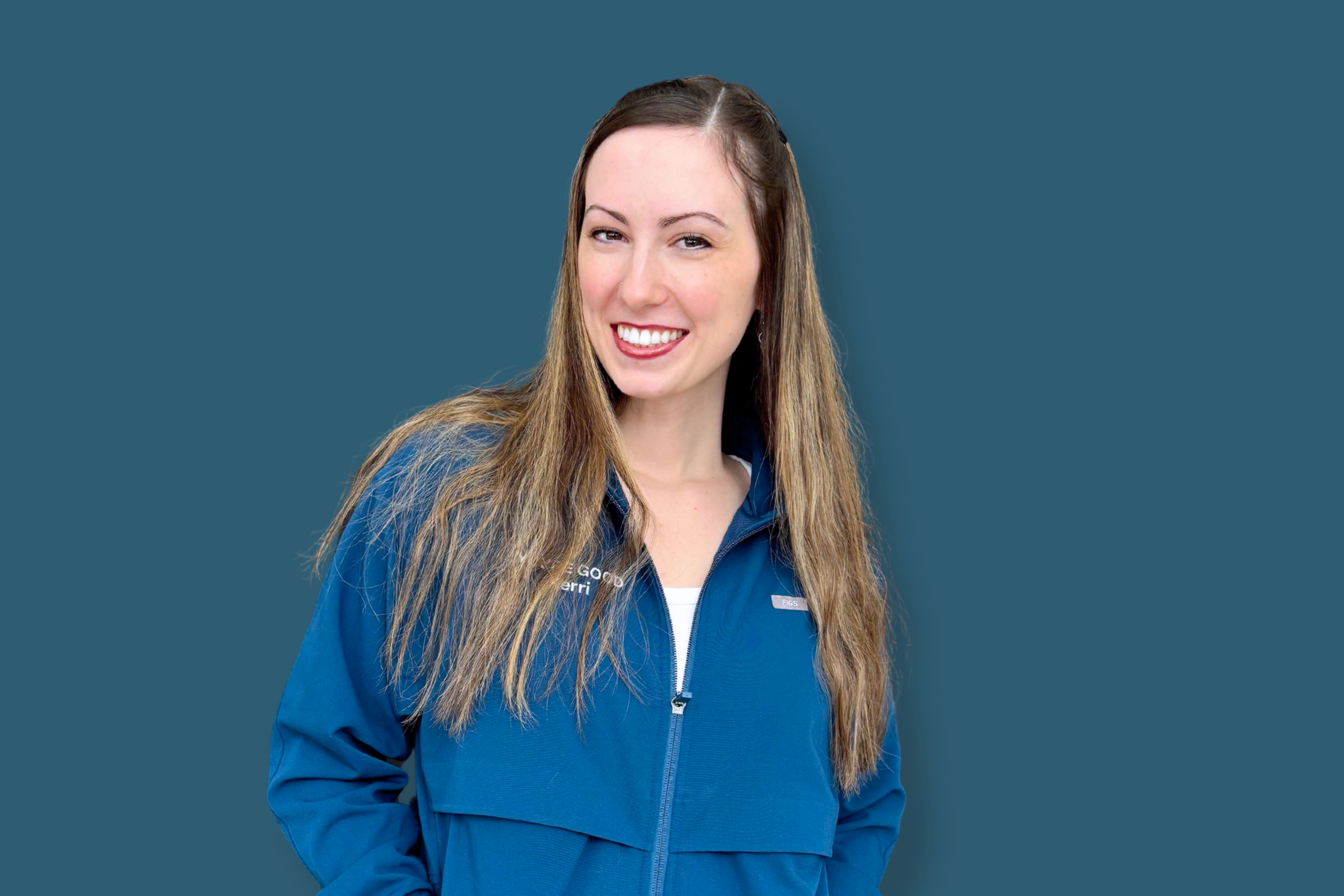 A woman with long brown hair wearing a blue zip-up jacket, smiling and looking at the camera against a solid blue background.