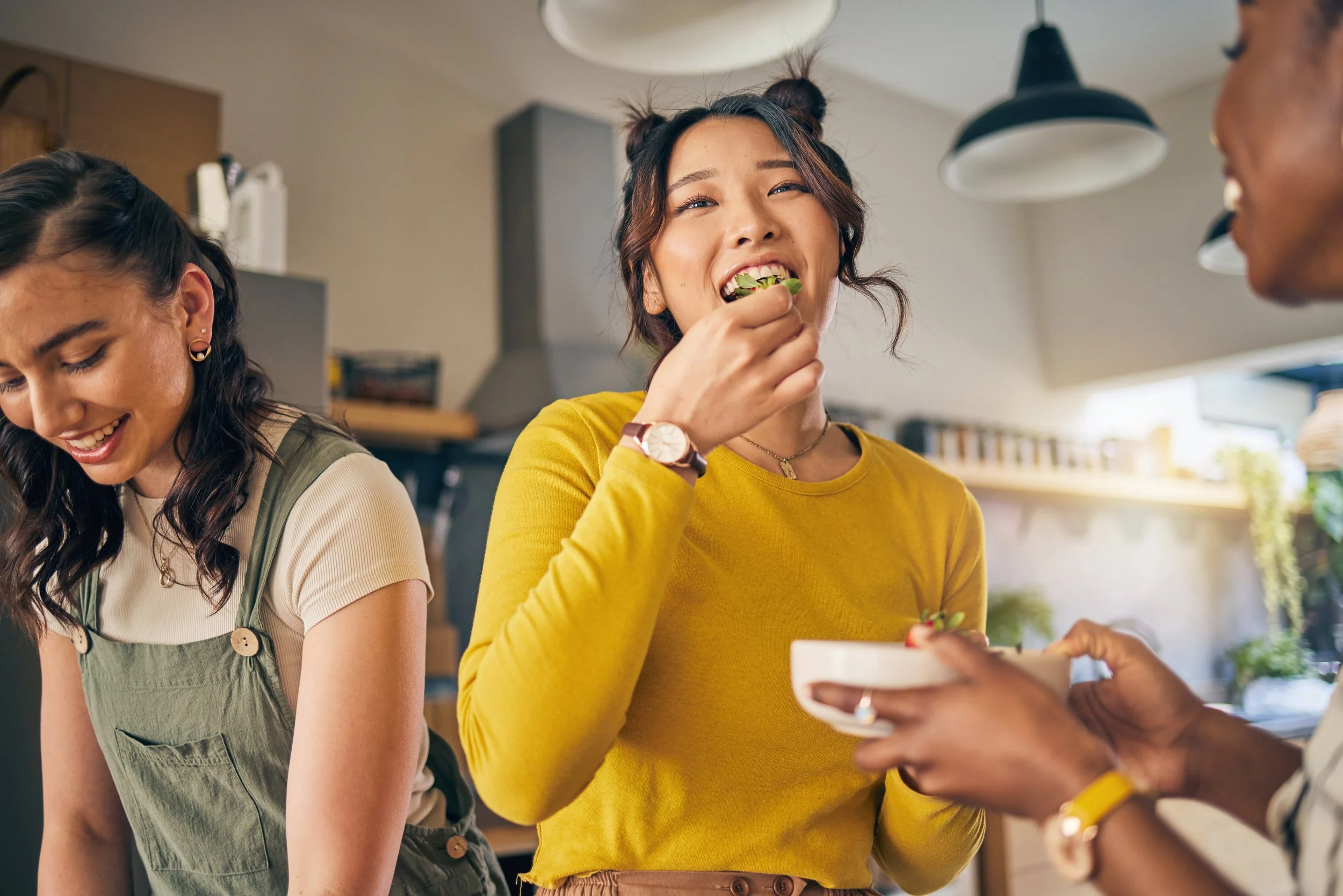 A group of young women enjoying a meal together, with one woman in a yellow top eating and smiling, while others are happily around her in a cozy kitchen setting.