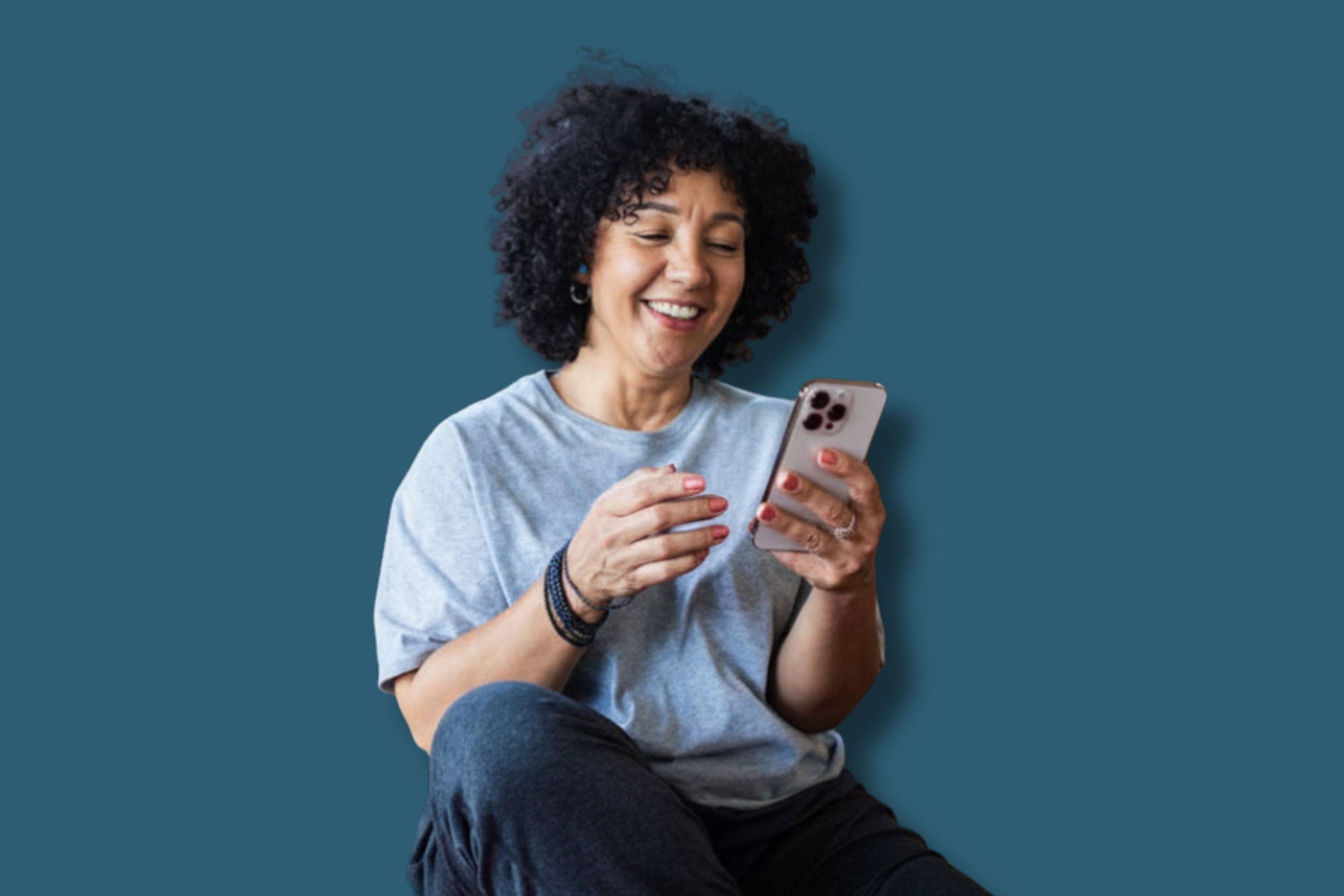 A woman with curly black hair smiling while looking at her smartphone, sitting against a blue background