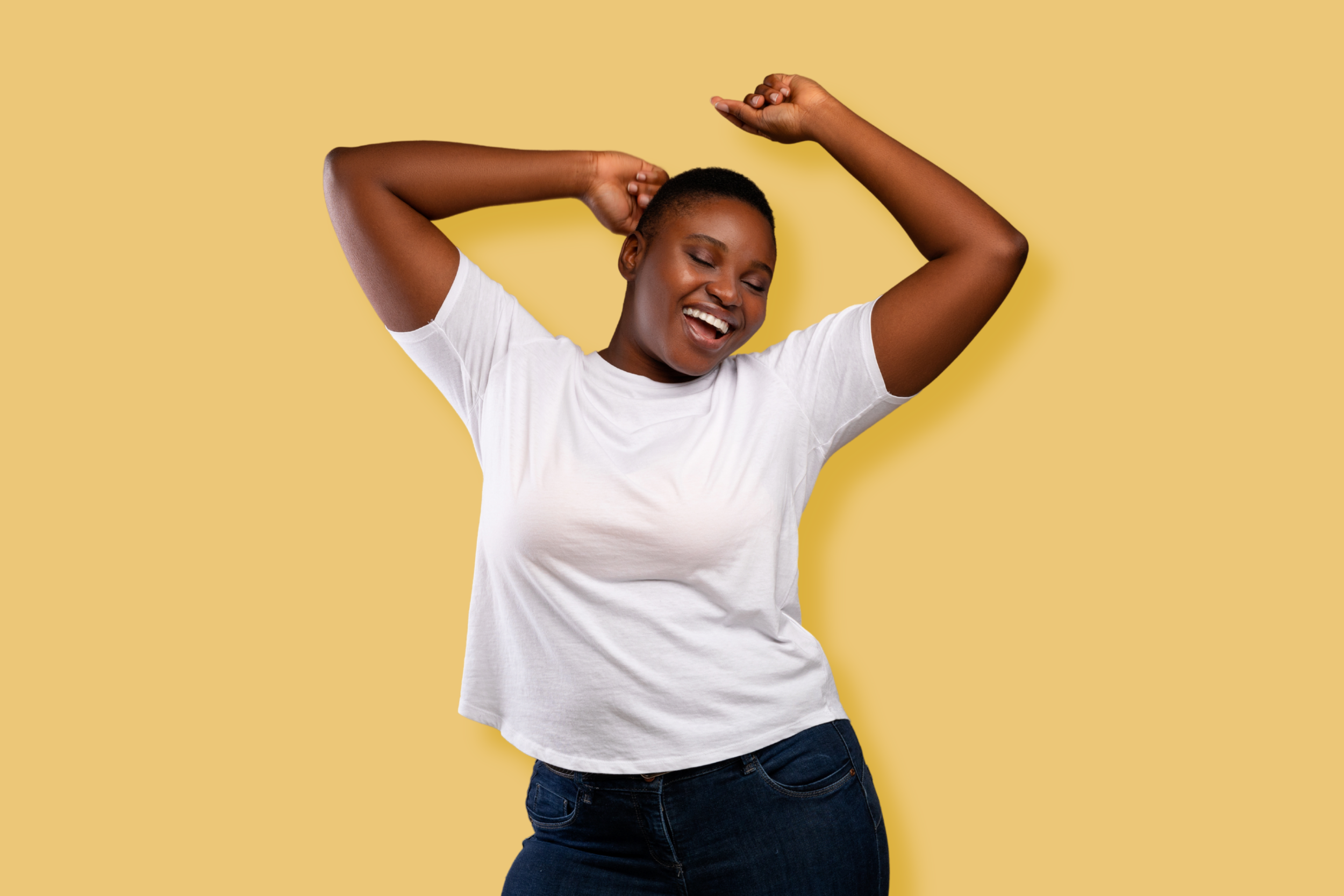 A woman with short hair, smiling with closed eyes, wearing a white t-shirt and blue jeans, standing against a yellow background with her arms raised and hands behind her head.