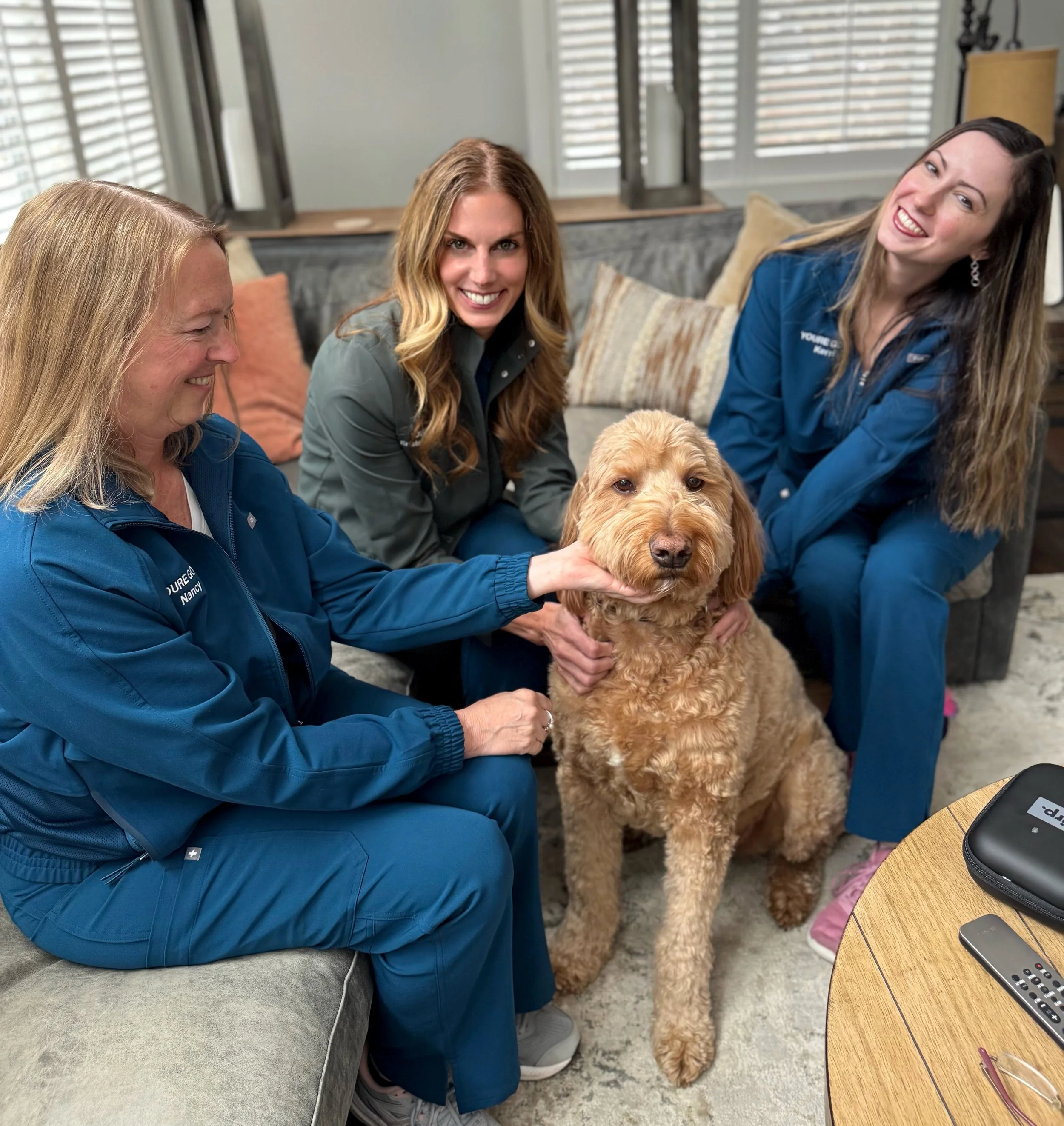 Three women in blue and gray uniforms smiling with a golden retriever dog inside a living room with a gray couch and window blinds.