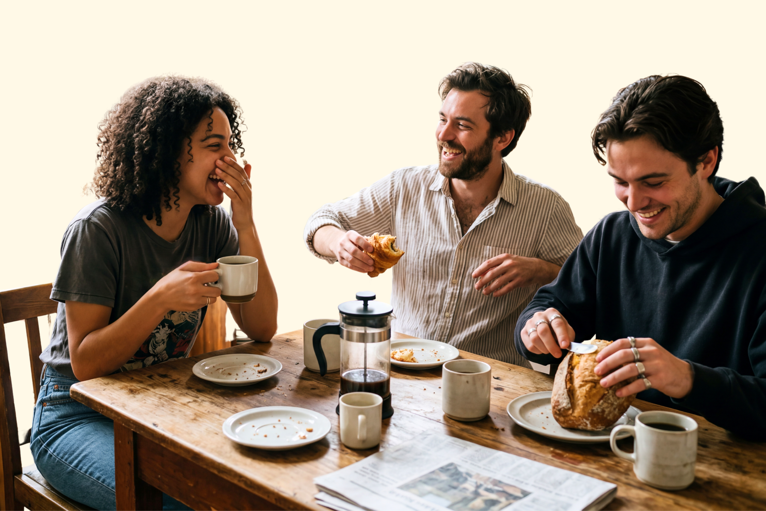 Three friends laughing and enjoying coffee and bread at a wooden table with plates, mugs, and a French press.