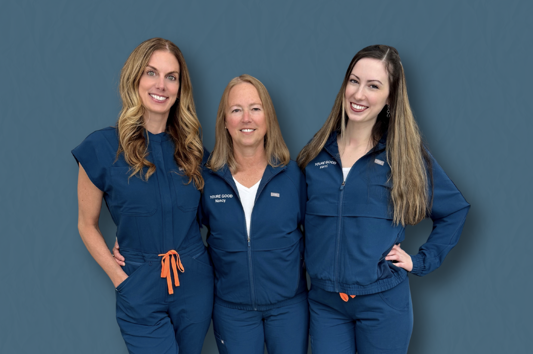 Three women in matching blue uniforms standing against a teal background. The woman on the left has long wavy blond hair, the woman in the middle has shoulder-length blond hair, and the woman on the right has long straight brown hair. They are smiling and looking at the camera.