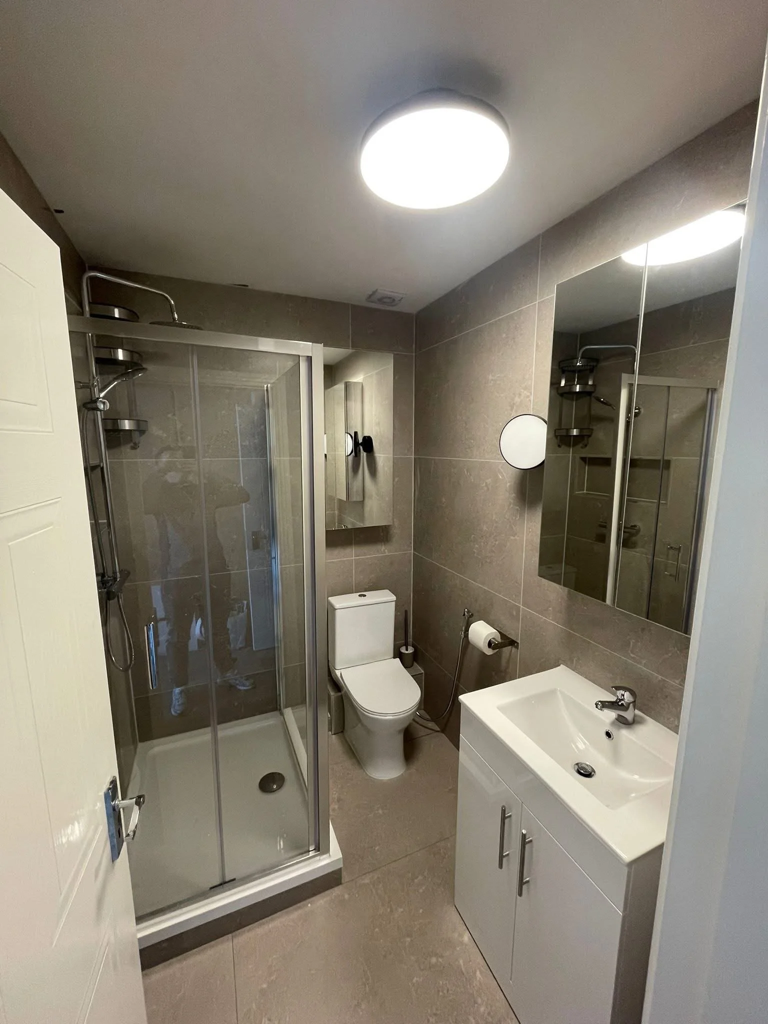 A modern bathroom with a shower stall, toilet, and a sink with cabinet, all against gray tiled walls and floor. There is a large circular ceiling light and a smaller wall mirror.