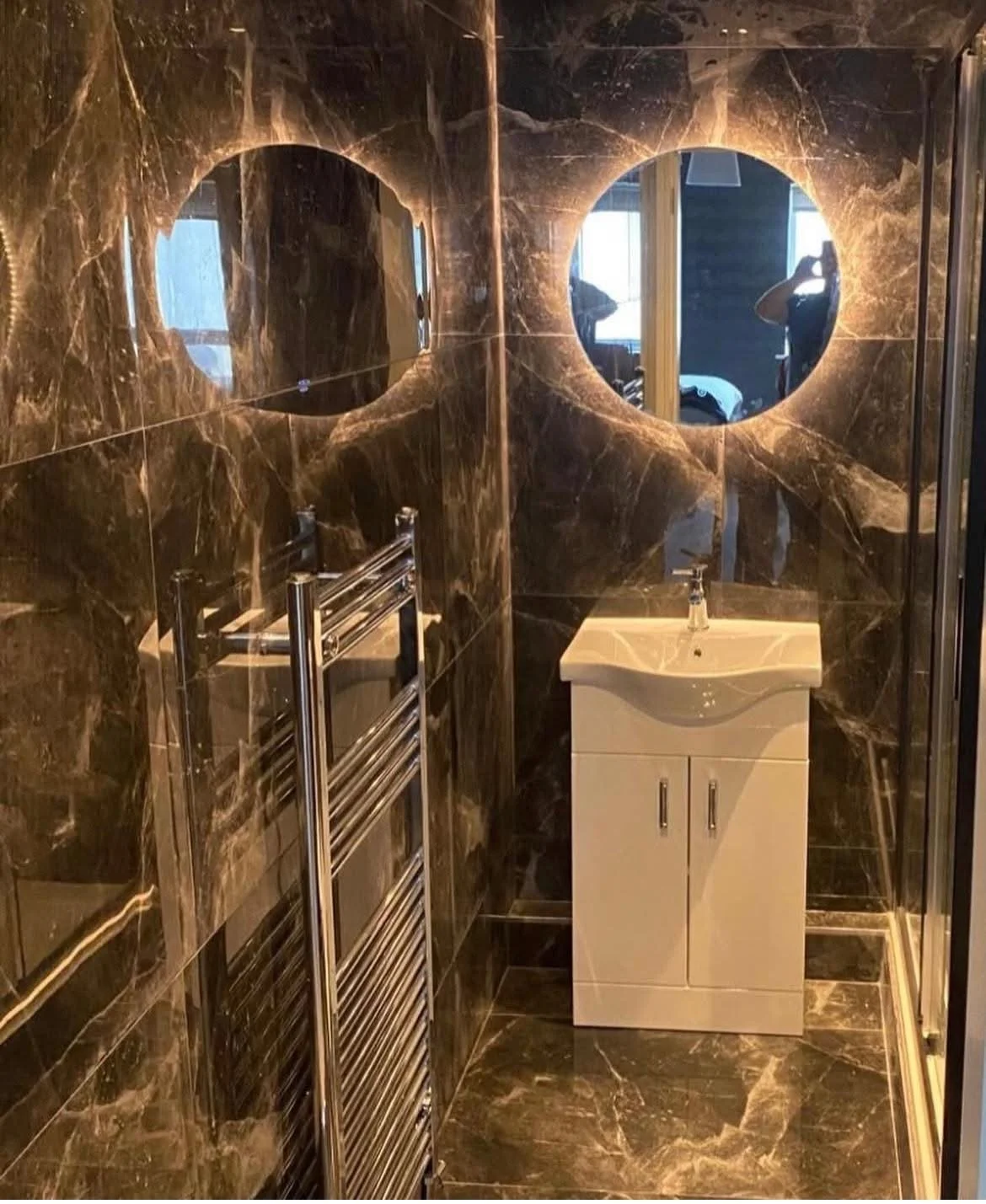 Small bathroom with dark brown marble walls and floor, a white sink with cabinet, a heated towel rail, and two round mirrors reflecting windows and a person taking the photo.