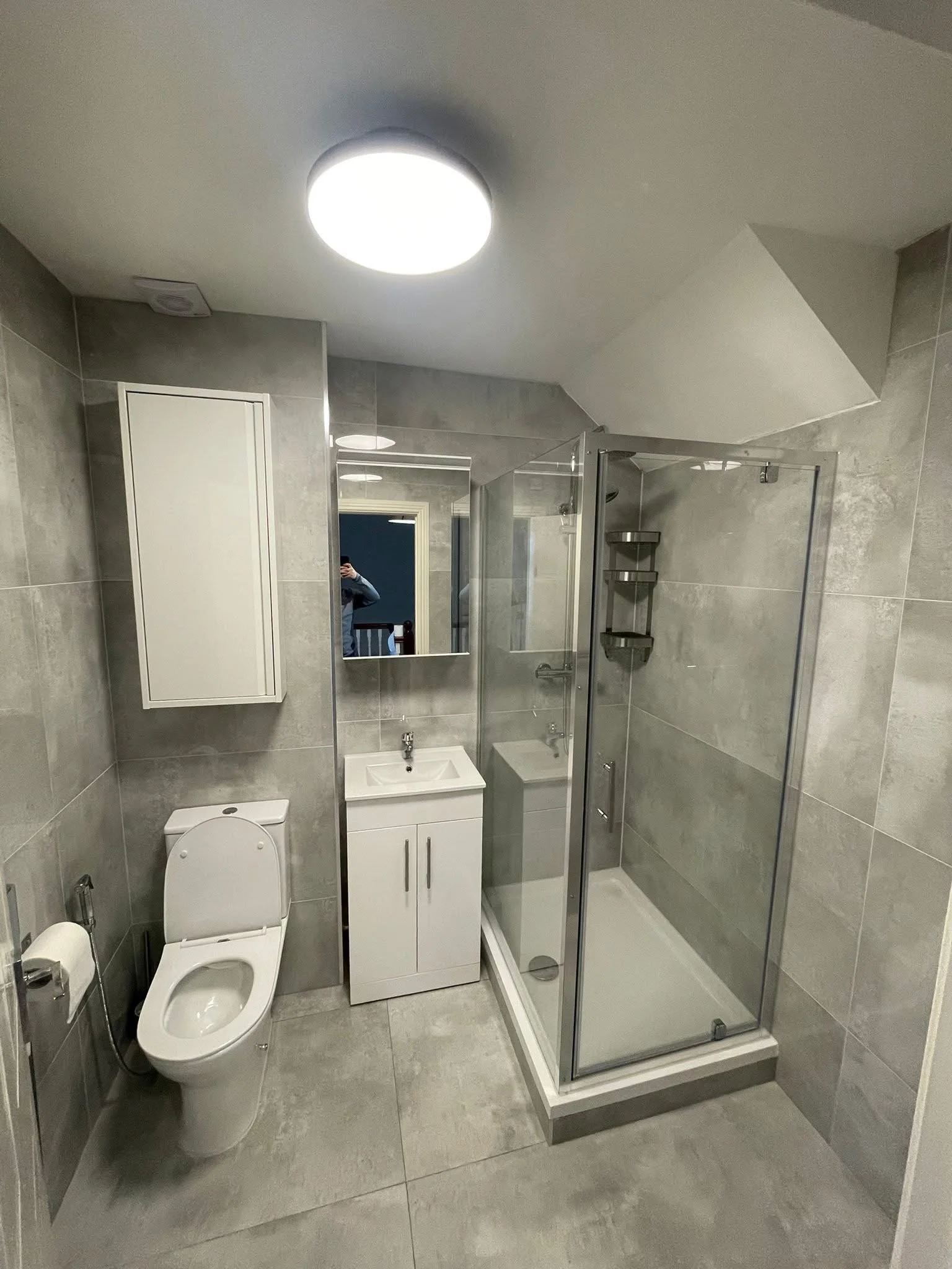 A small modern bathroom with gray tiled walls and floor, a toilet, a small white vanity with a sink, a mirror, a glass-enclosed shower with a shower bench and shelves, and a wall-mounted cabinet.