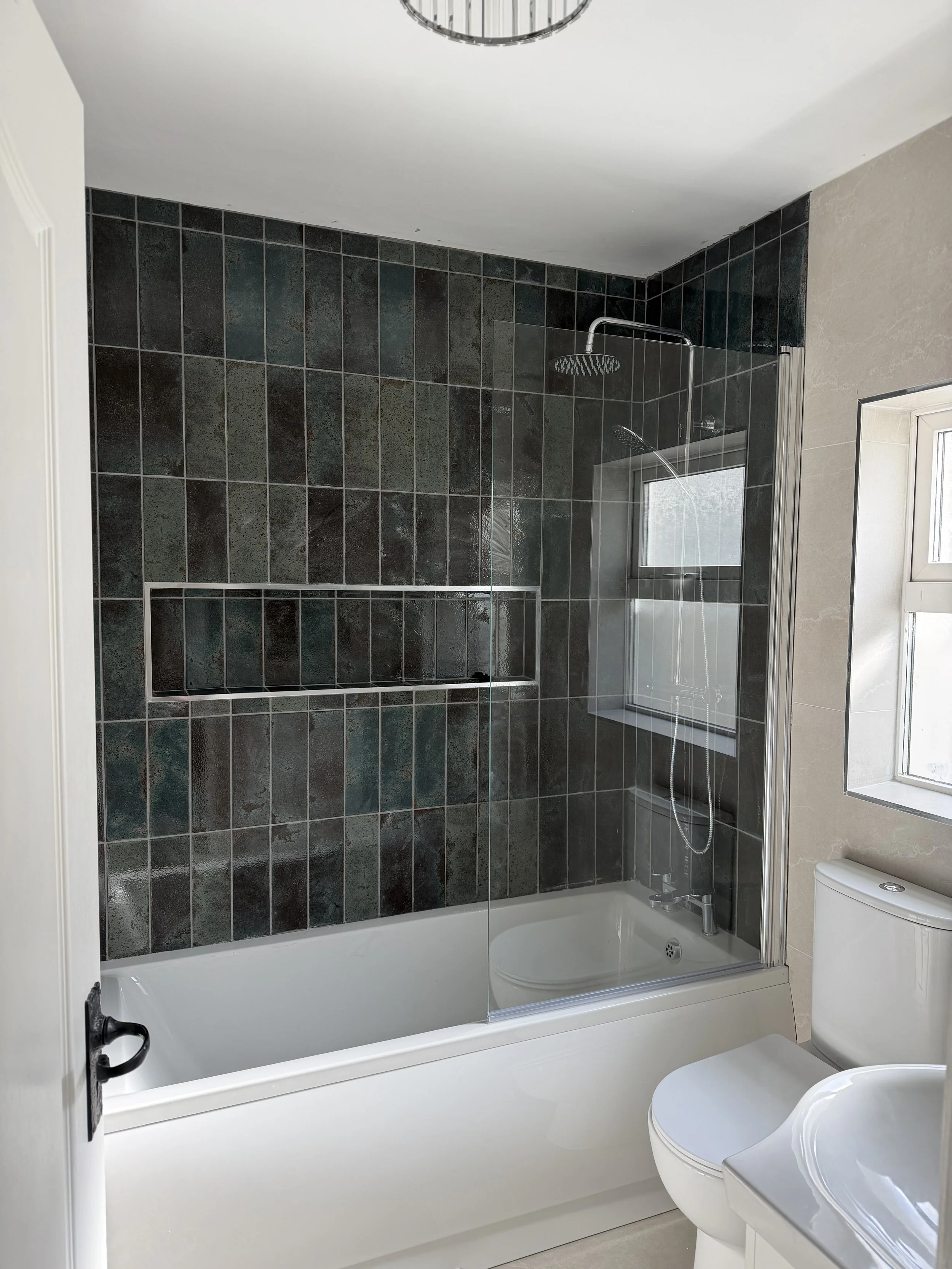 Bathroom with bathtub, glass shower enclosure, dark tiled wall, window, toilet, and sink.