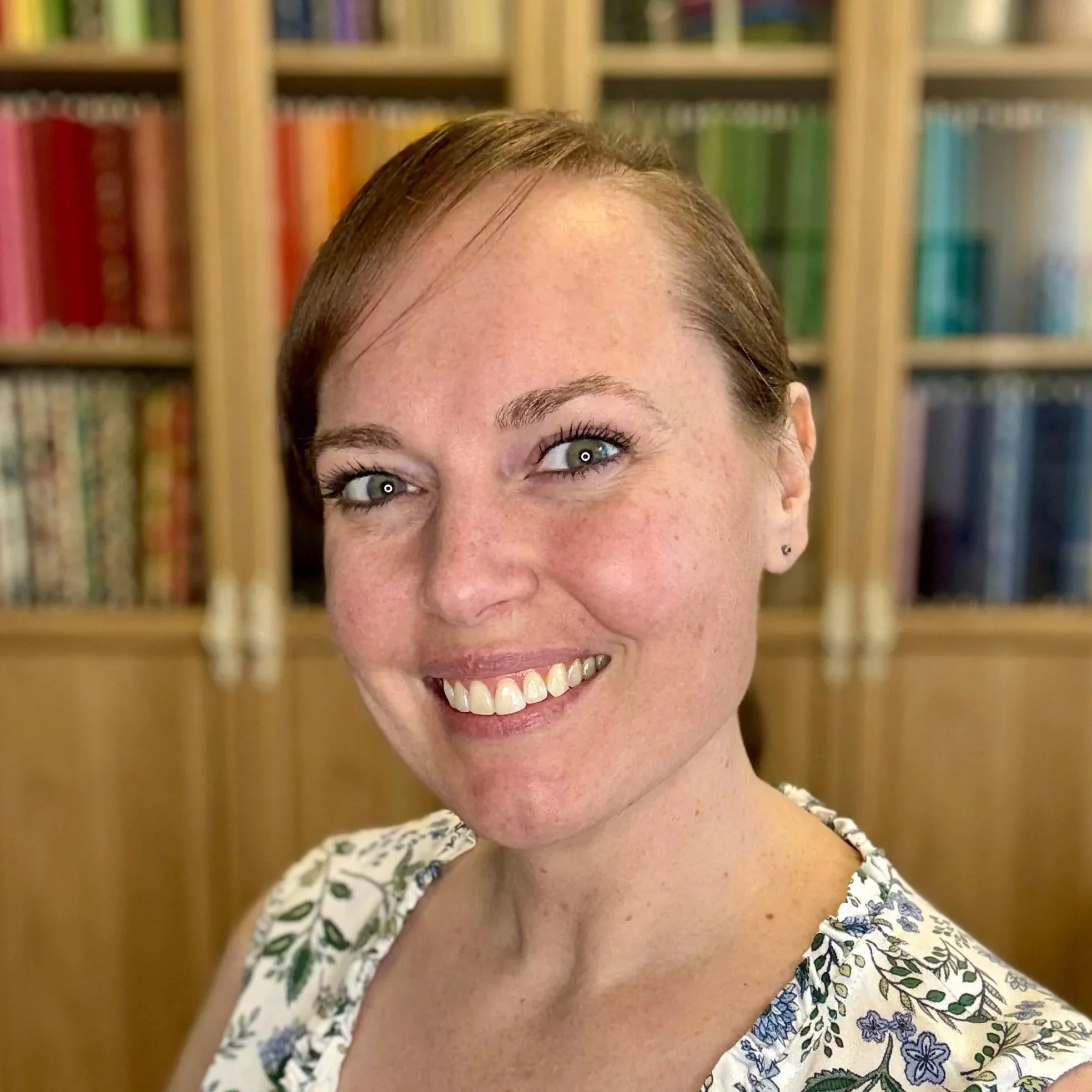 A smiling woman with brown hair and freckles, wearing a floral-patterned top, standing in front of a bookshelf with colorful fabrics.
