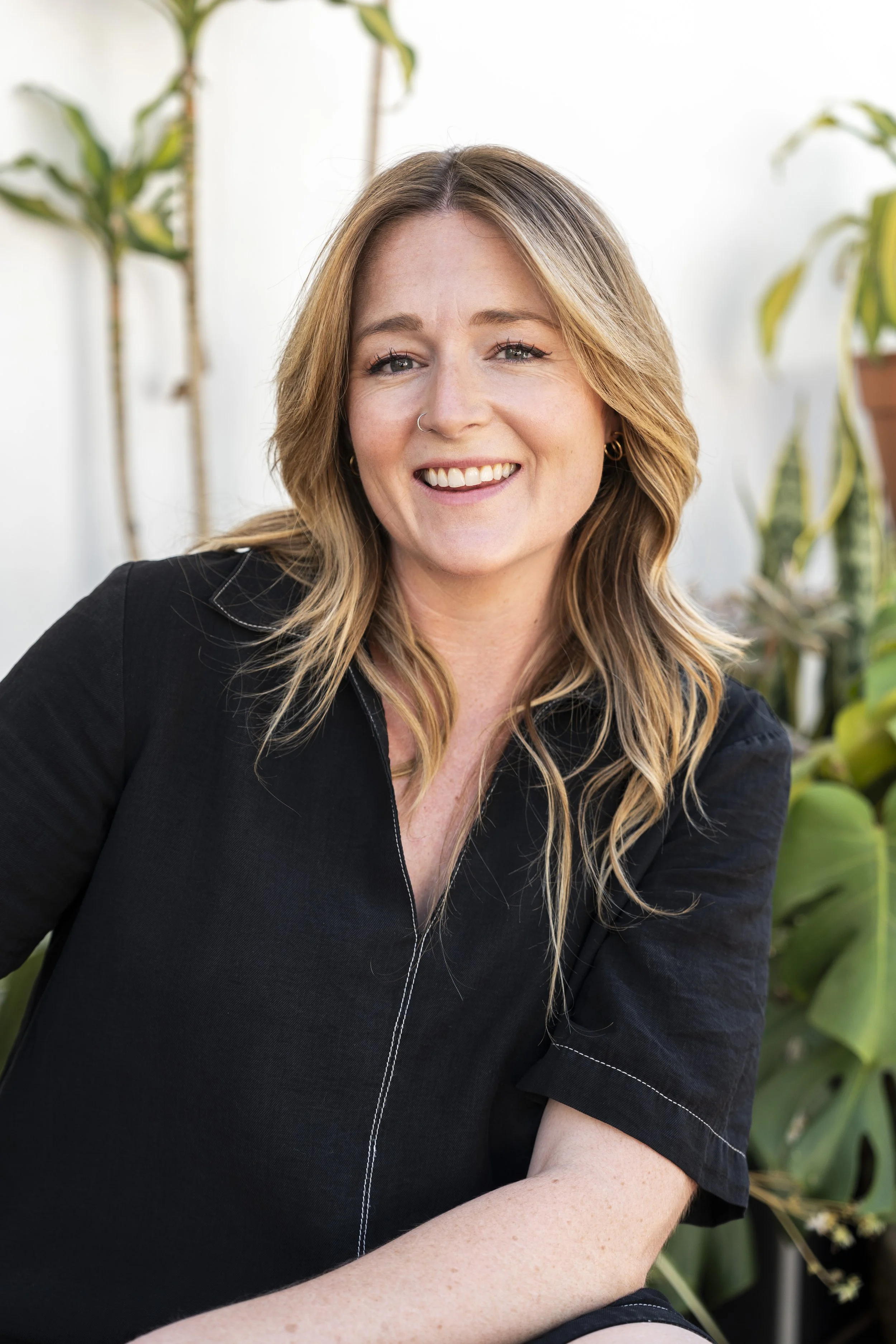 Pheobe Hanley, a woman with blonde hair and a nose piercing smiling, wearing a black shirt, sitting outdoors with green plants in the background.