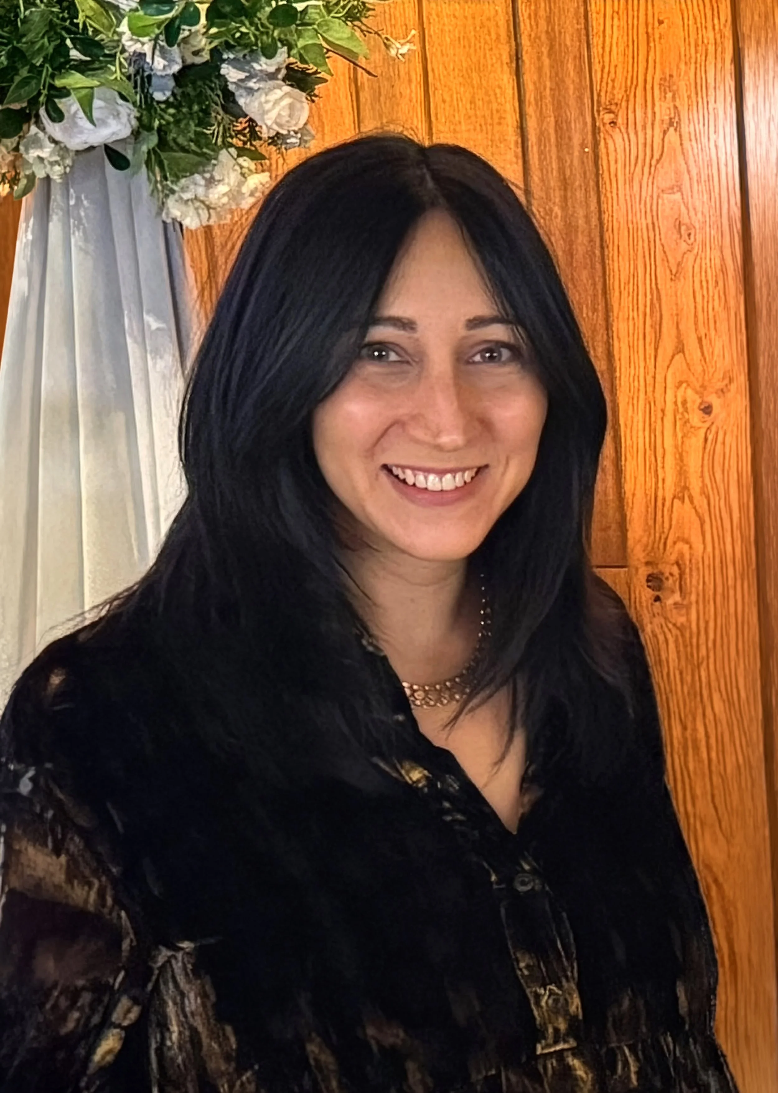 Kirsten Donovan, a woman with long black hair smiling, wearing a dark velvet dress with gold details, standing in front of a wooden-paneled wall with floral decorations to her left.