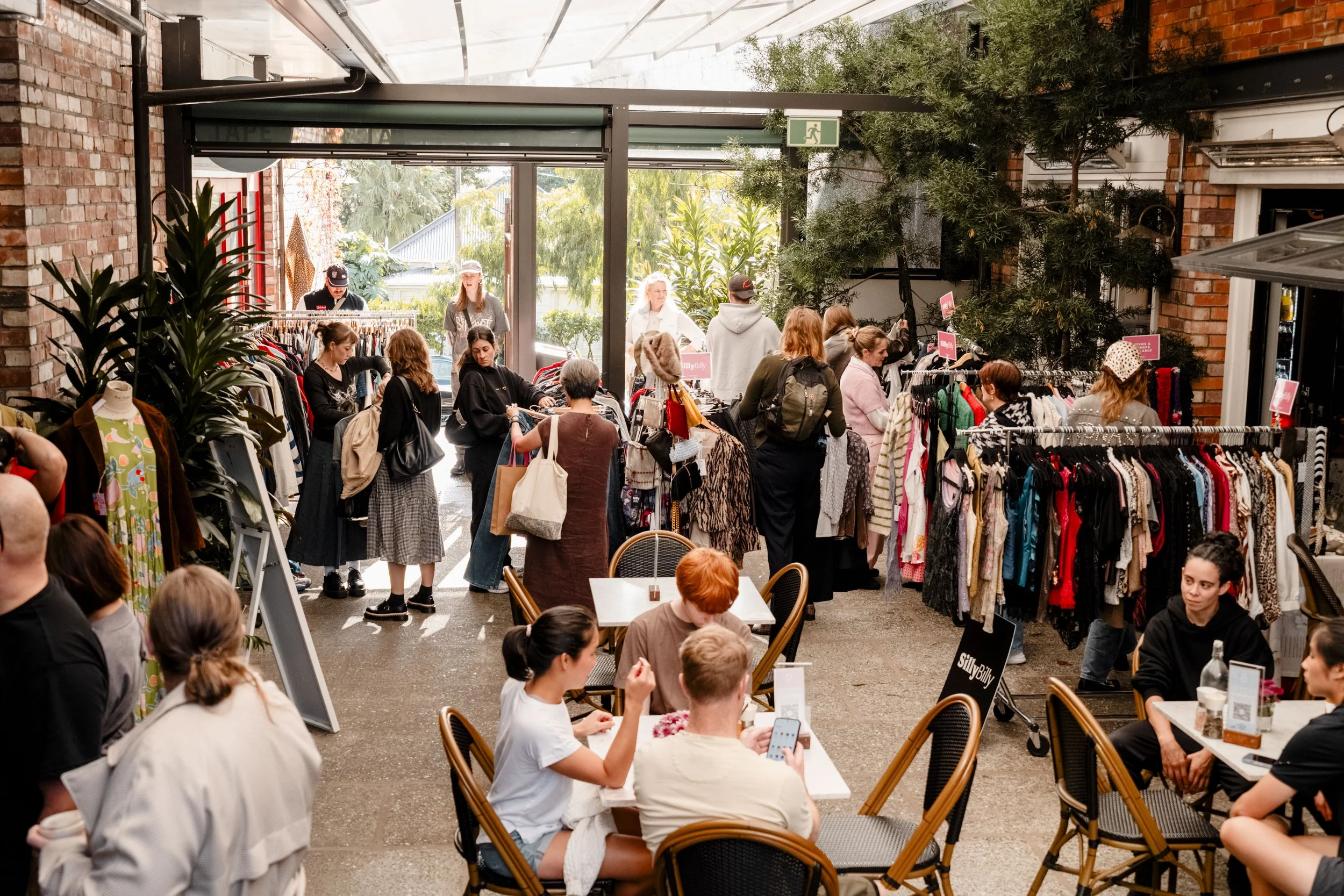 People browsing clothing racks at an indoor flea market or thrift store, with some sitting at tables and others shopping.