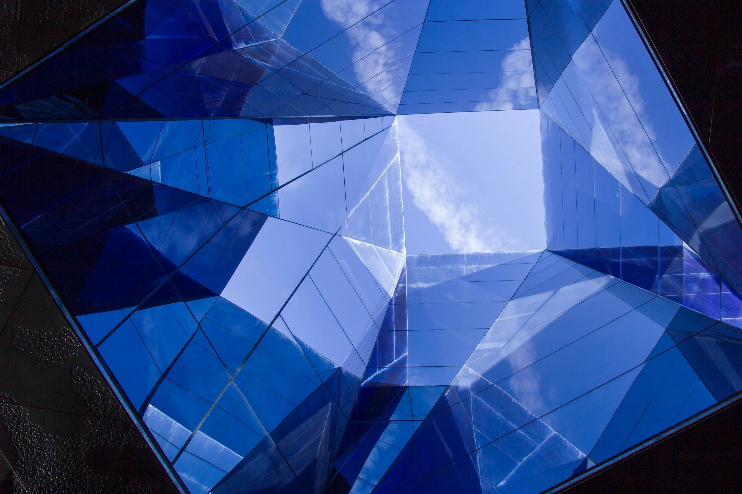 Reflection of blue sky and clouds on the glass facade of a modern building, viewed from the bottom looking up.