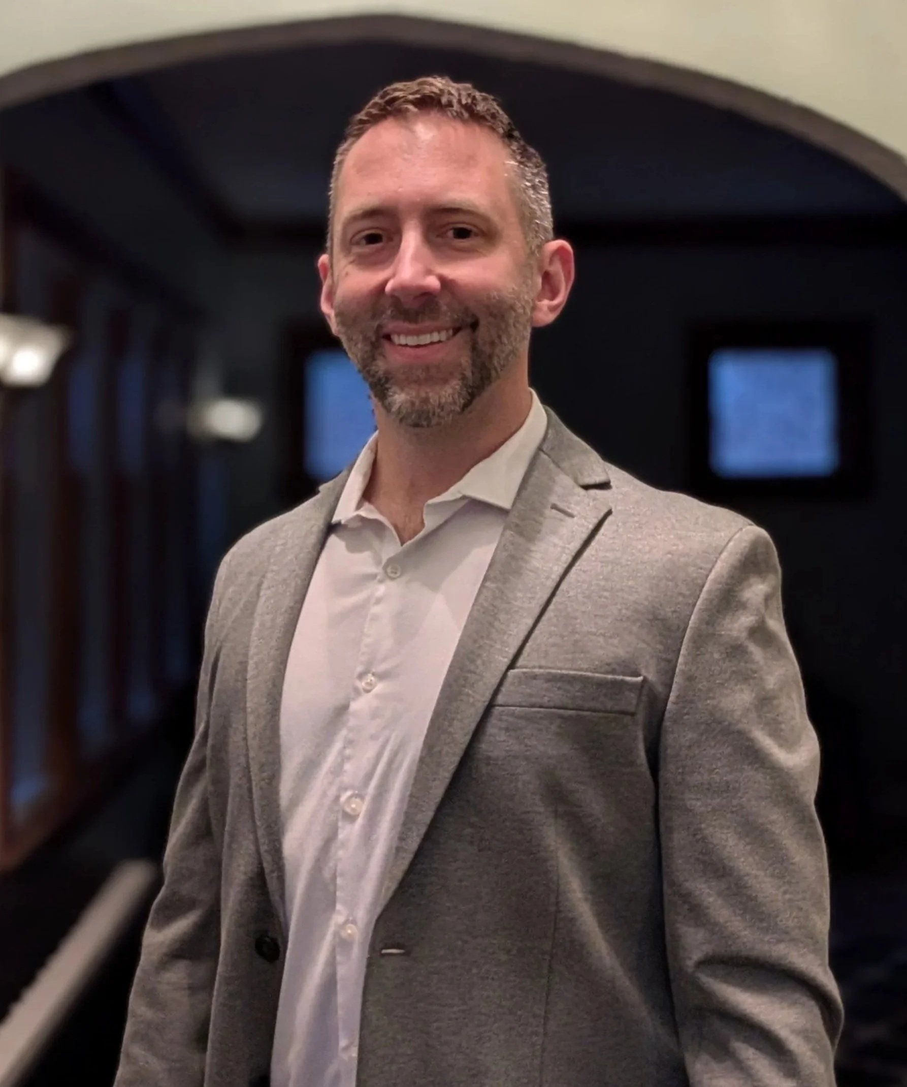 A smiling man with a beard in a gray suit and white shirt standing indoors near windows.