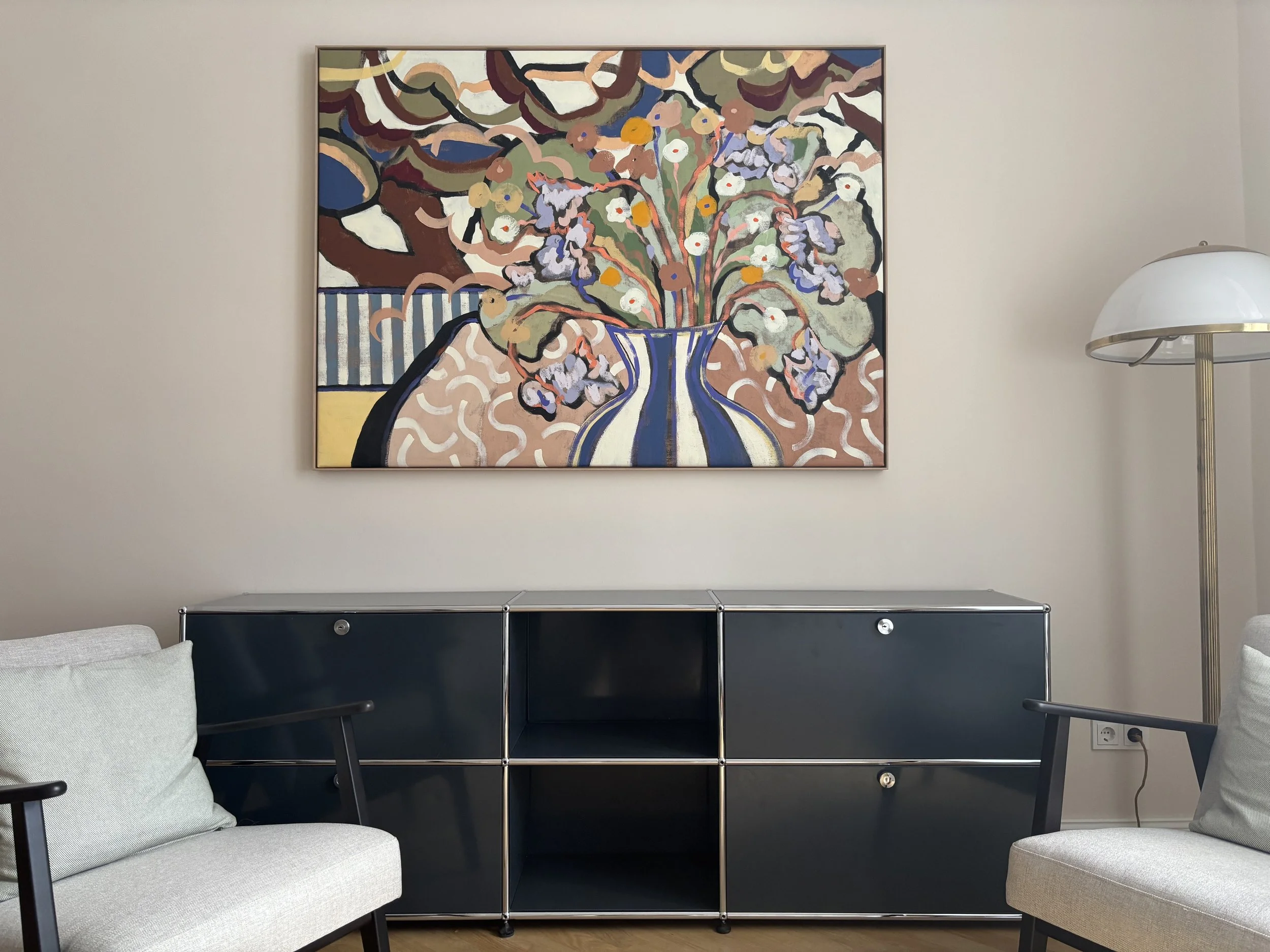 Ein stilvolles psychiatrisch bzw. psychotherapeutisches Praxiszimmer mit einem bunten abstrakten Gemälde an der Wand, zwei weiße Sessel und ein dunkler Schrank. Ein Stehlampe steht an der rechten Seite.
