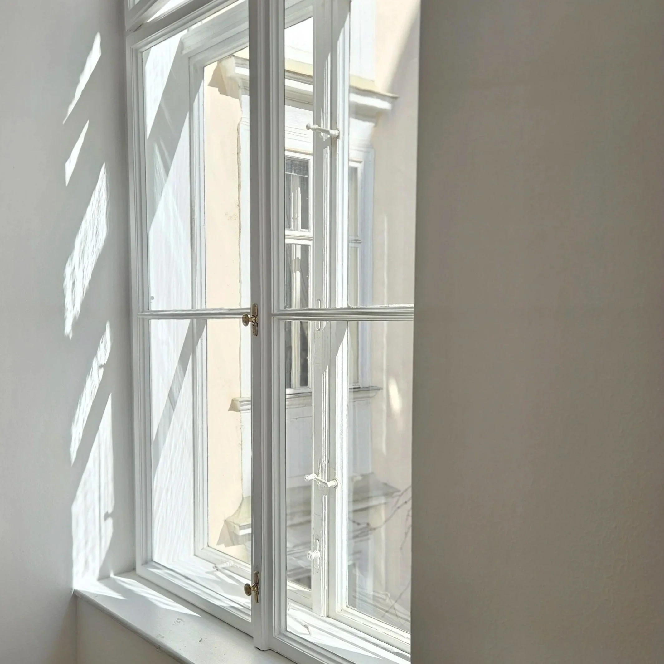 Ein weißes Fenster mit mehreren Glasfenstern, durch die Sonnenlicht in den Raum scheint und Schatten an die Wand werfen.