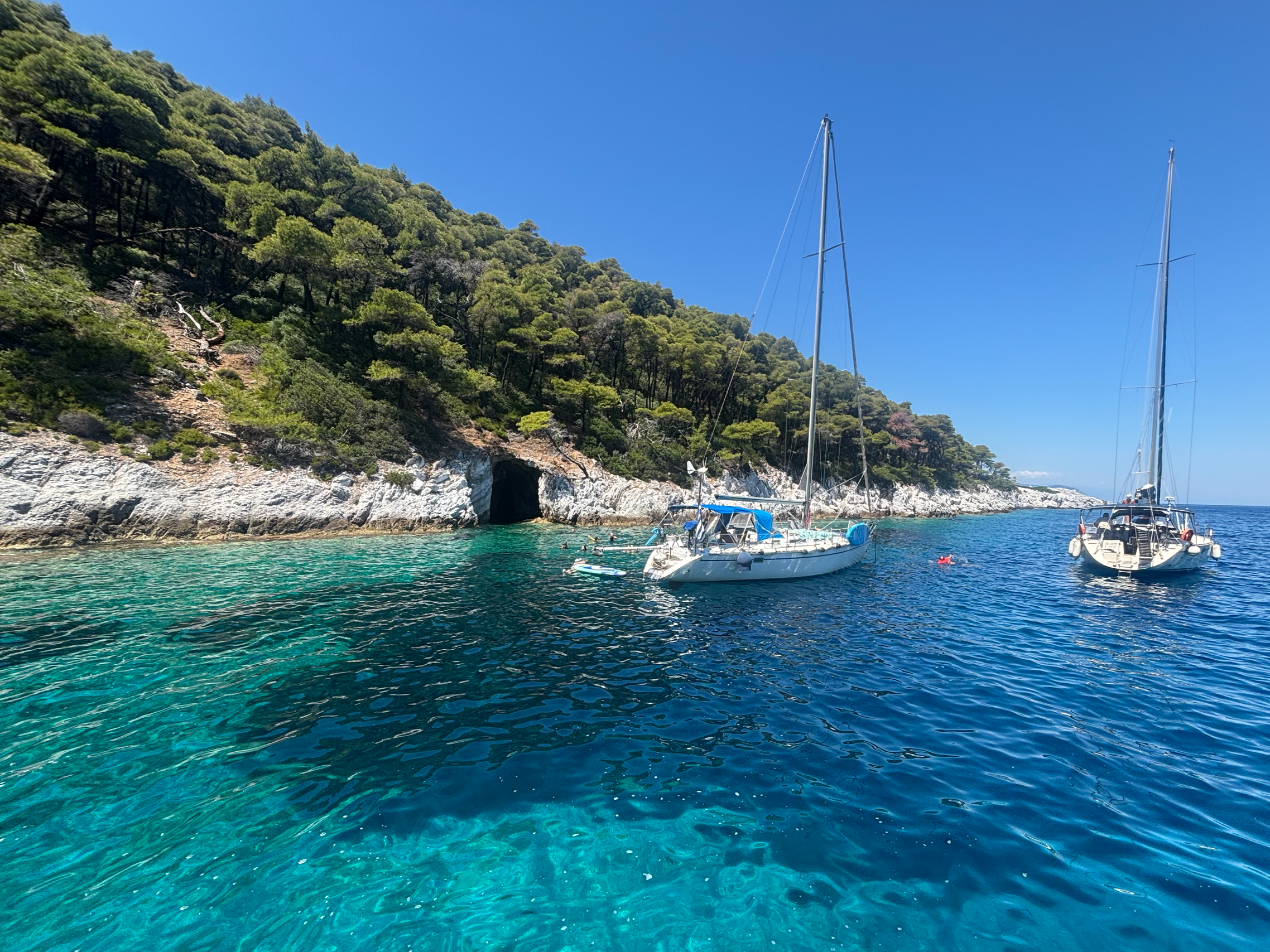 A view of boats anchored near a rocky shoreline with a green hillside and a cave, under a clear blue sky.