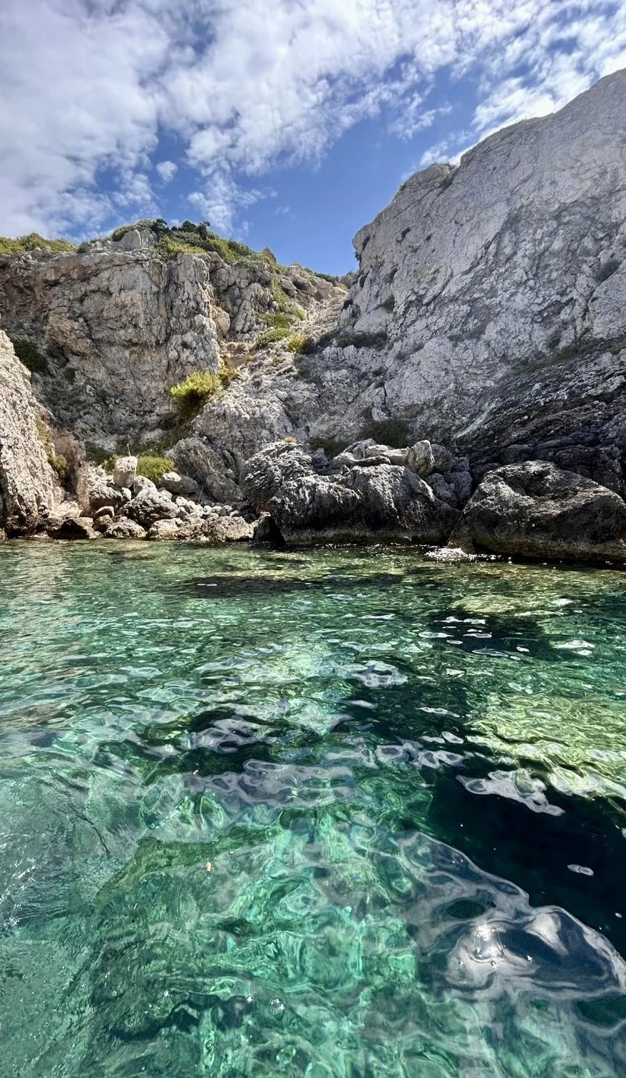 A clear, greenish-blue body of water surrounded by rocky cliffs and large boulders under a partly cloudy sky.