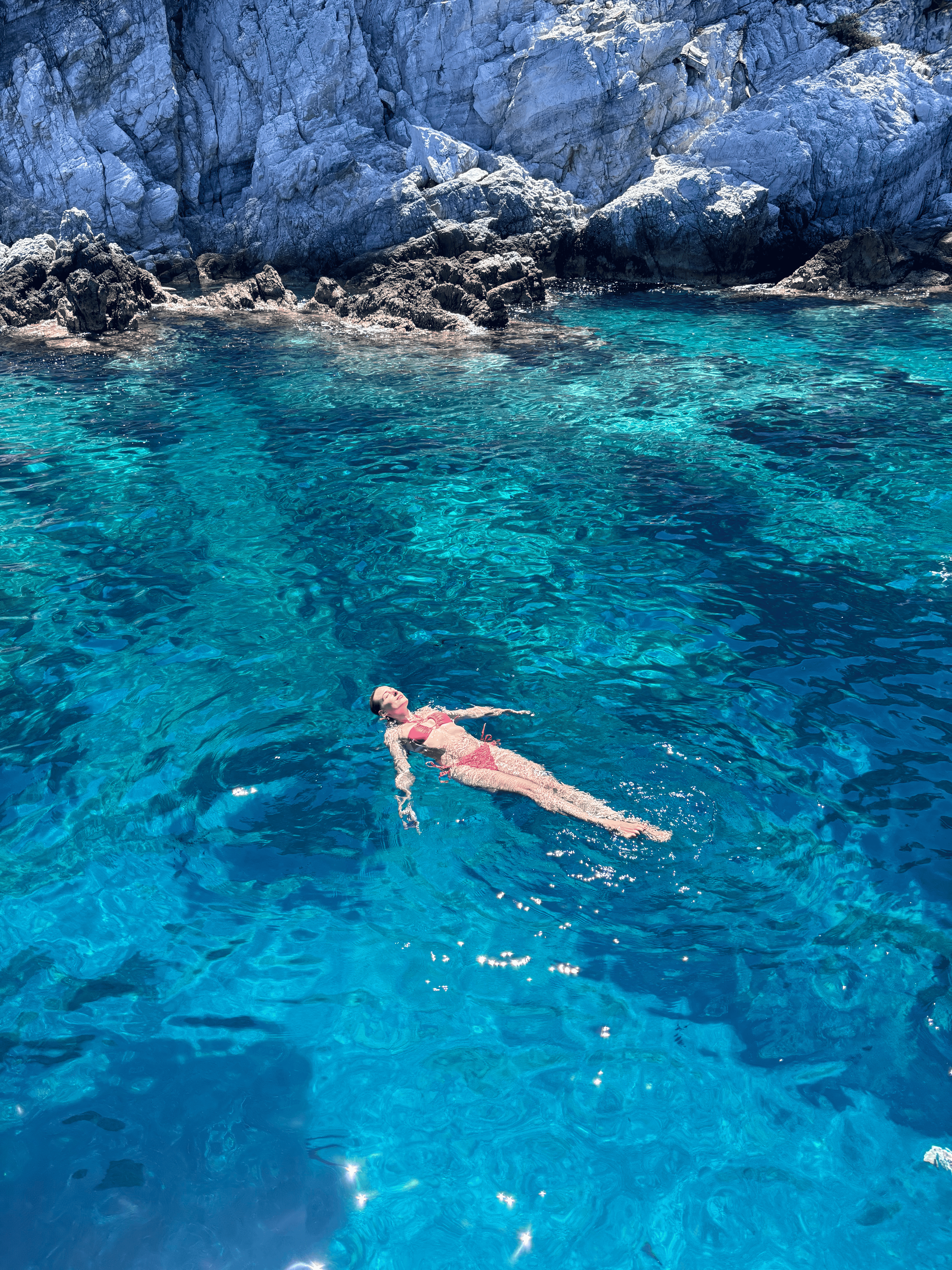 A person in a pink swimsuit floating on their back in clear blue water near rocky cliffs.