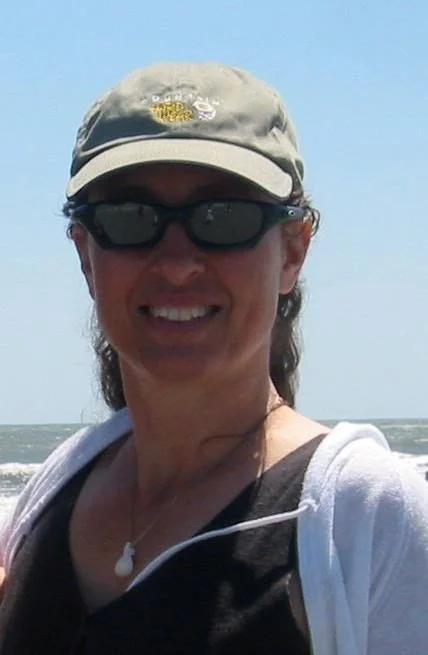 Woman wearing a gray cap, dark sunglasses, and a white soft sweater outdoors, with the ocean in the background.