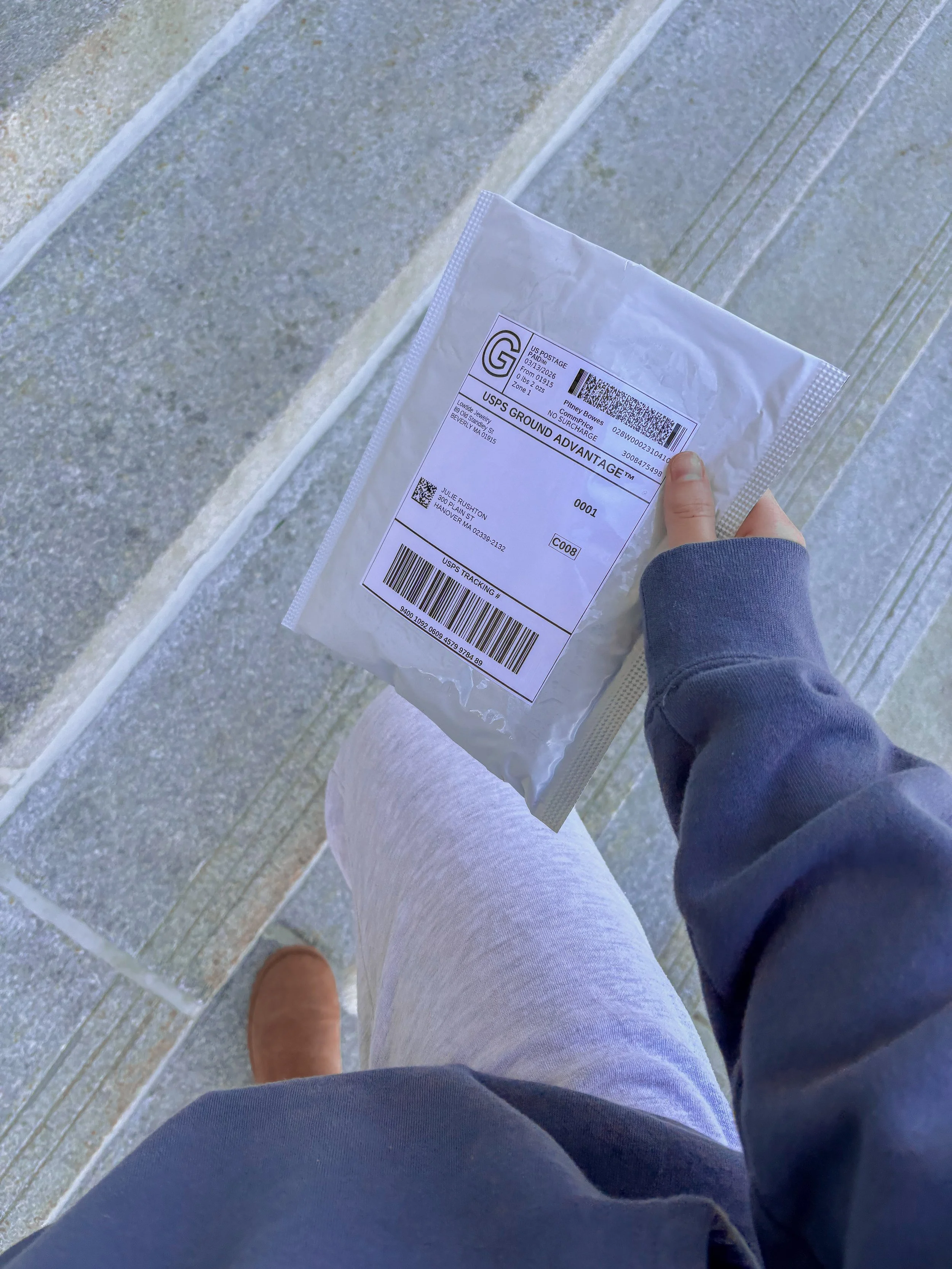 Person standing on tile floor holding a USPS shipping package, seen from above, wearing a navy hoodie, beige pants, and brown shoes.