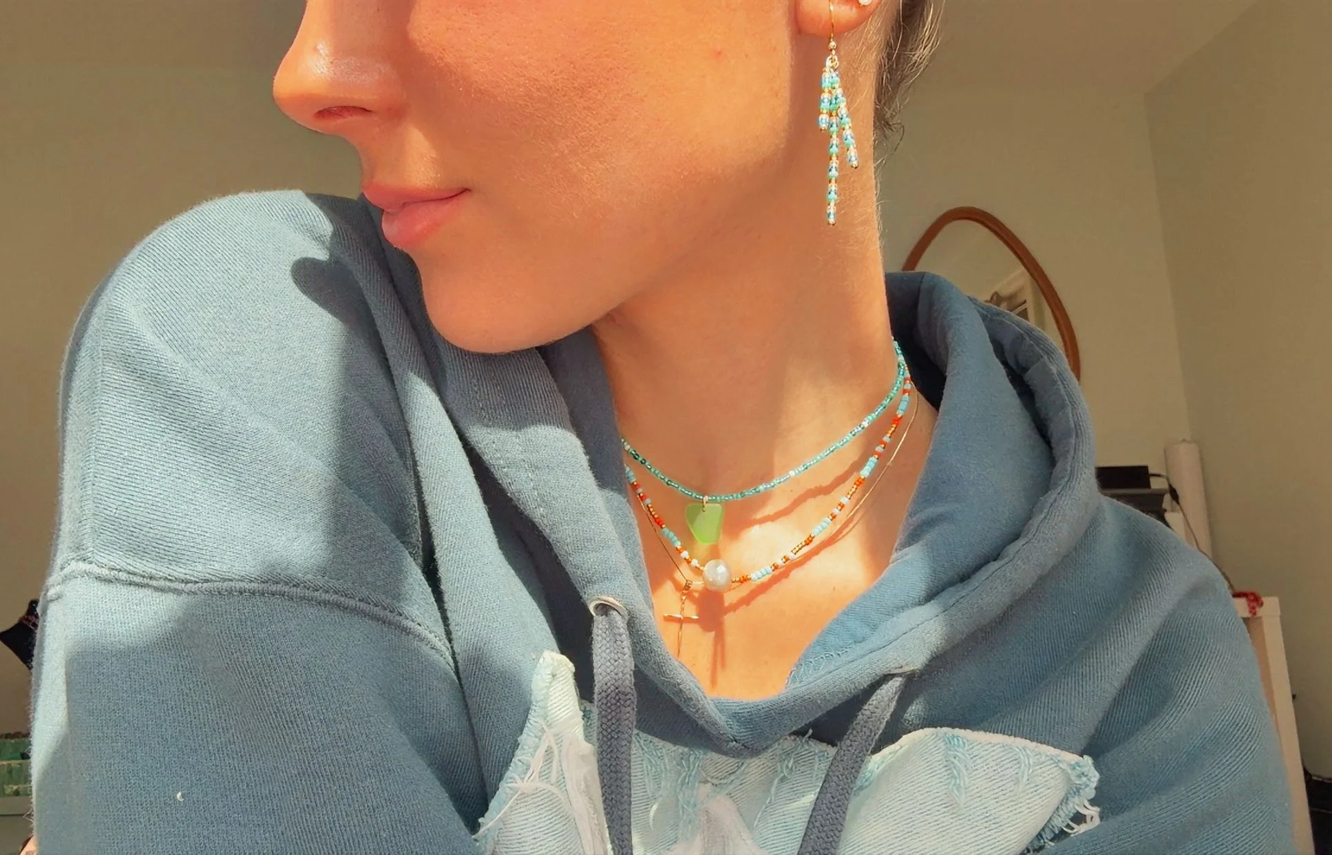 Close-up of a woman wearing colorful layered necklaces, dangling earrings, and a blue hoodie.