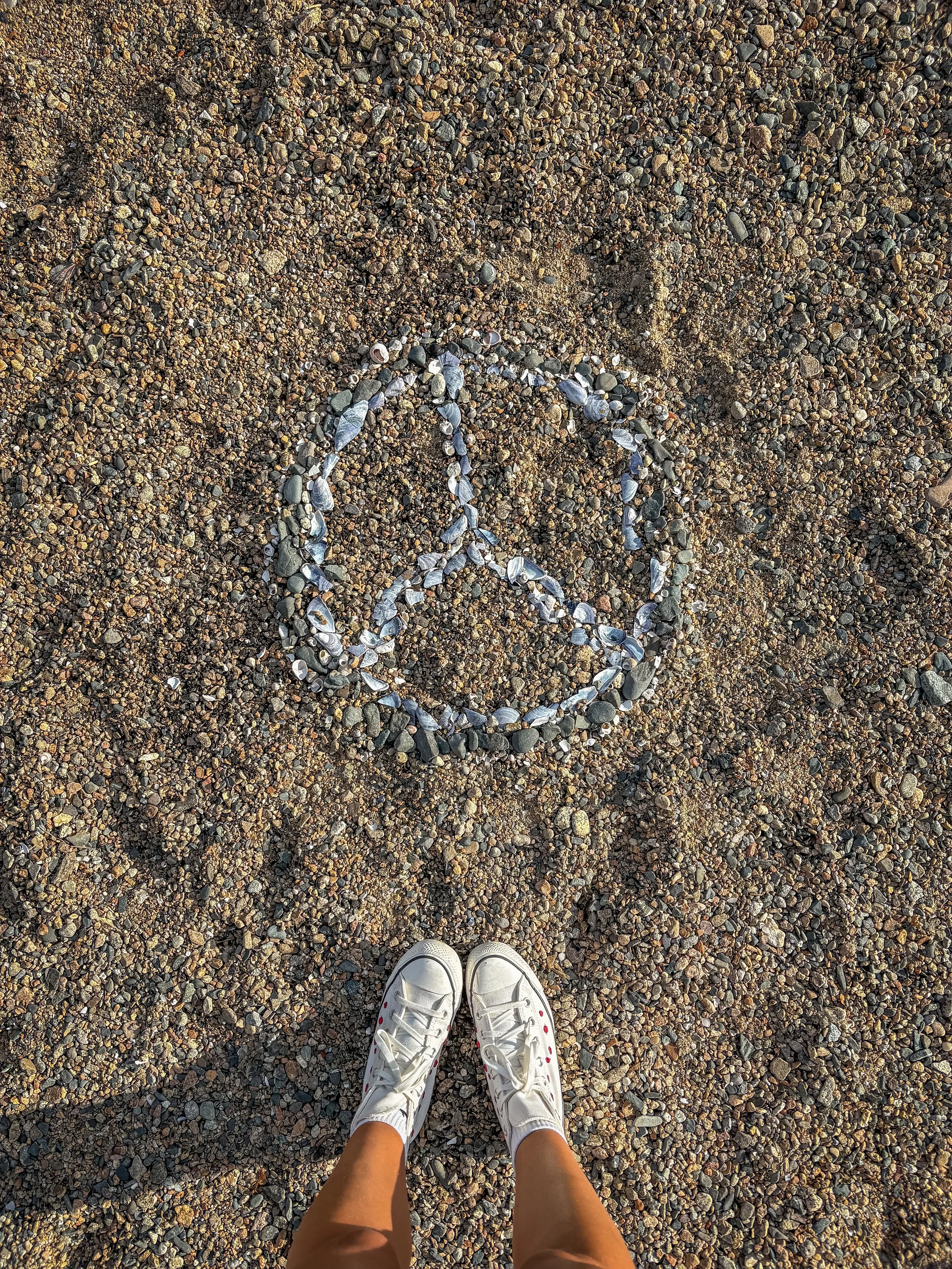 Person standing on gravel beach, wearing white sneakers, looking down at a peace symbol drawn with seashells on the ground.