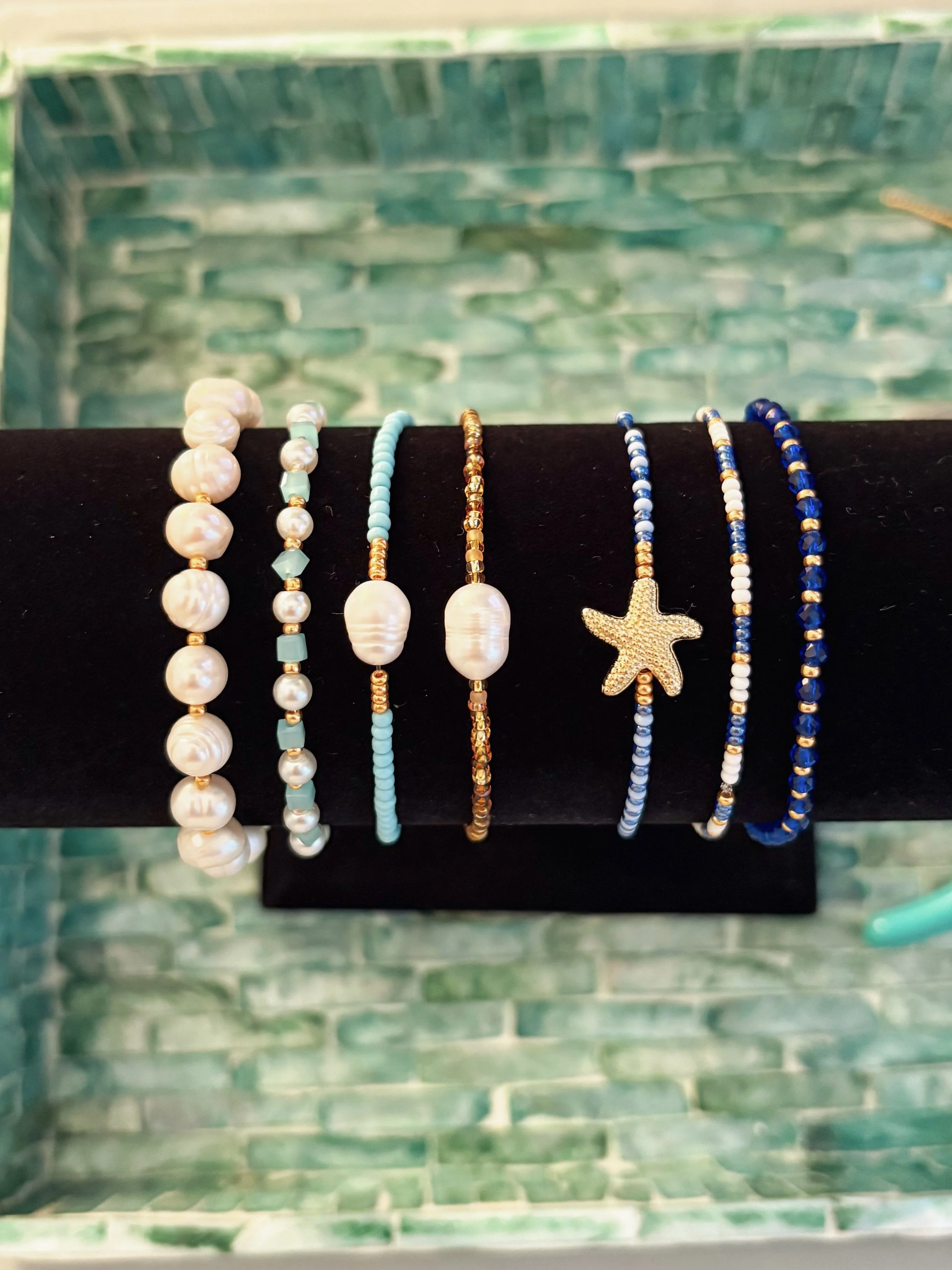 A display of seven different beaded bracelets with various colors and designs on a black stand, against a green tiled wall background.