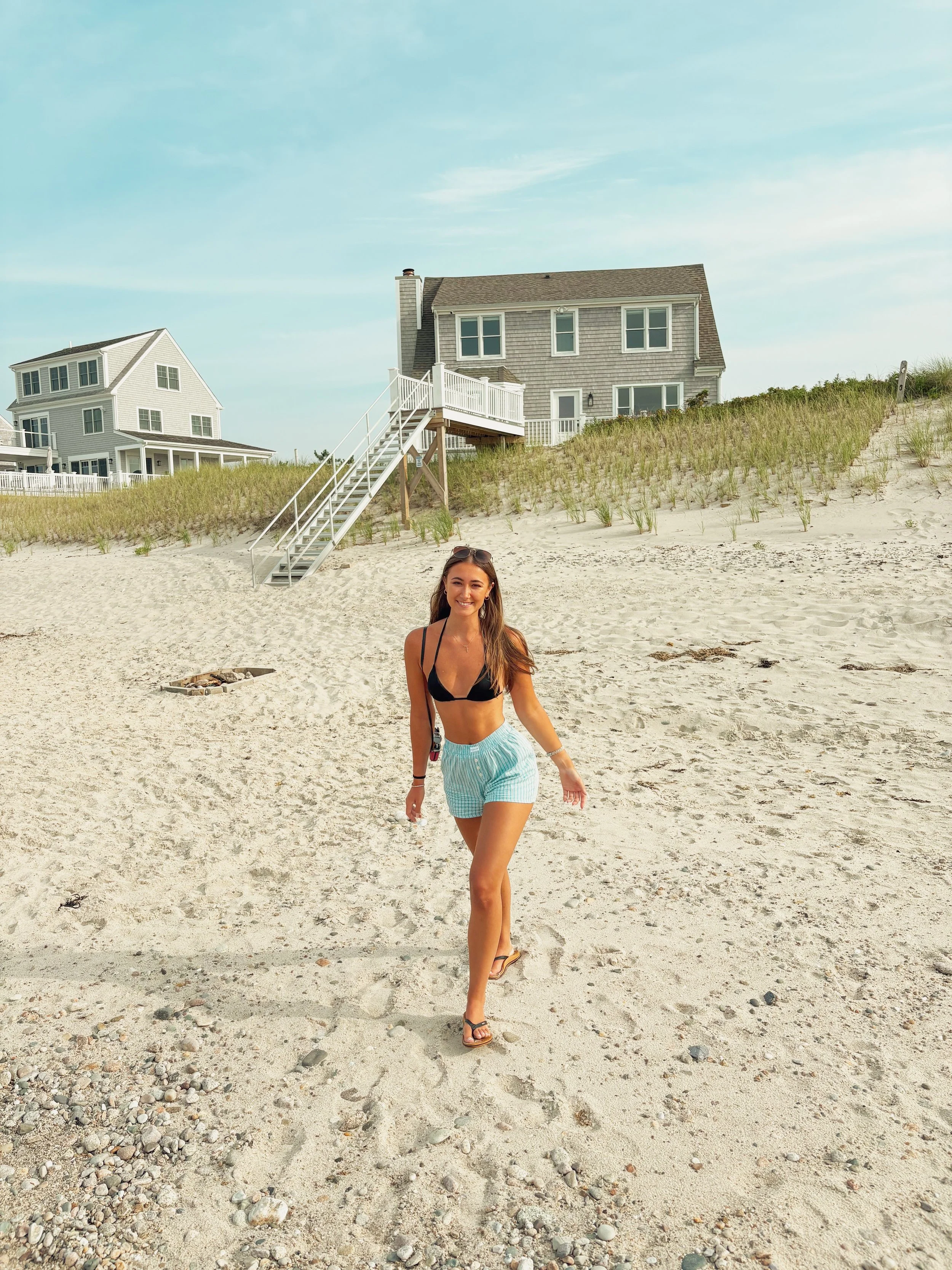Young woman in black bikini top and striped shorts walking on sandy beach with houses on dune in background.