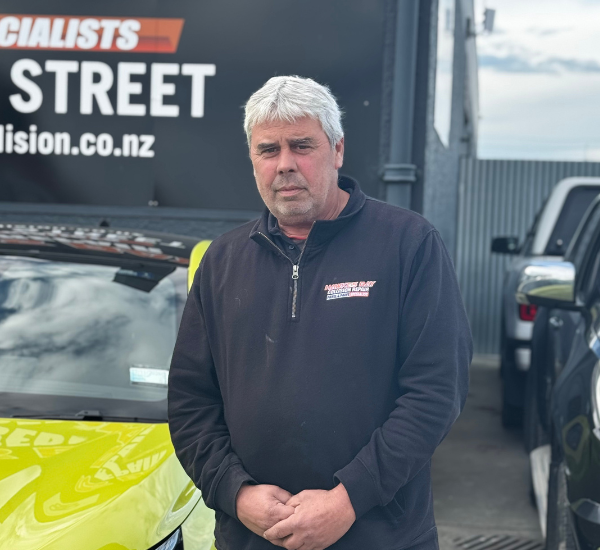 A middle-aged man with gray hair, wearing a black jacket with a logo on it, standing outdoors in front of a yellow car and a black sign with white and red lettering.