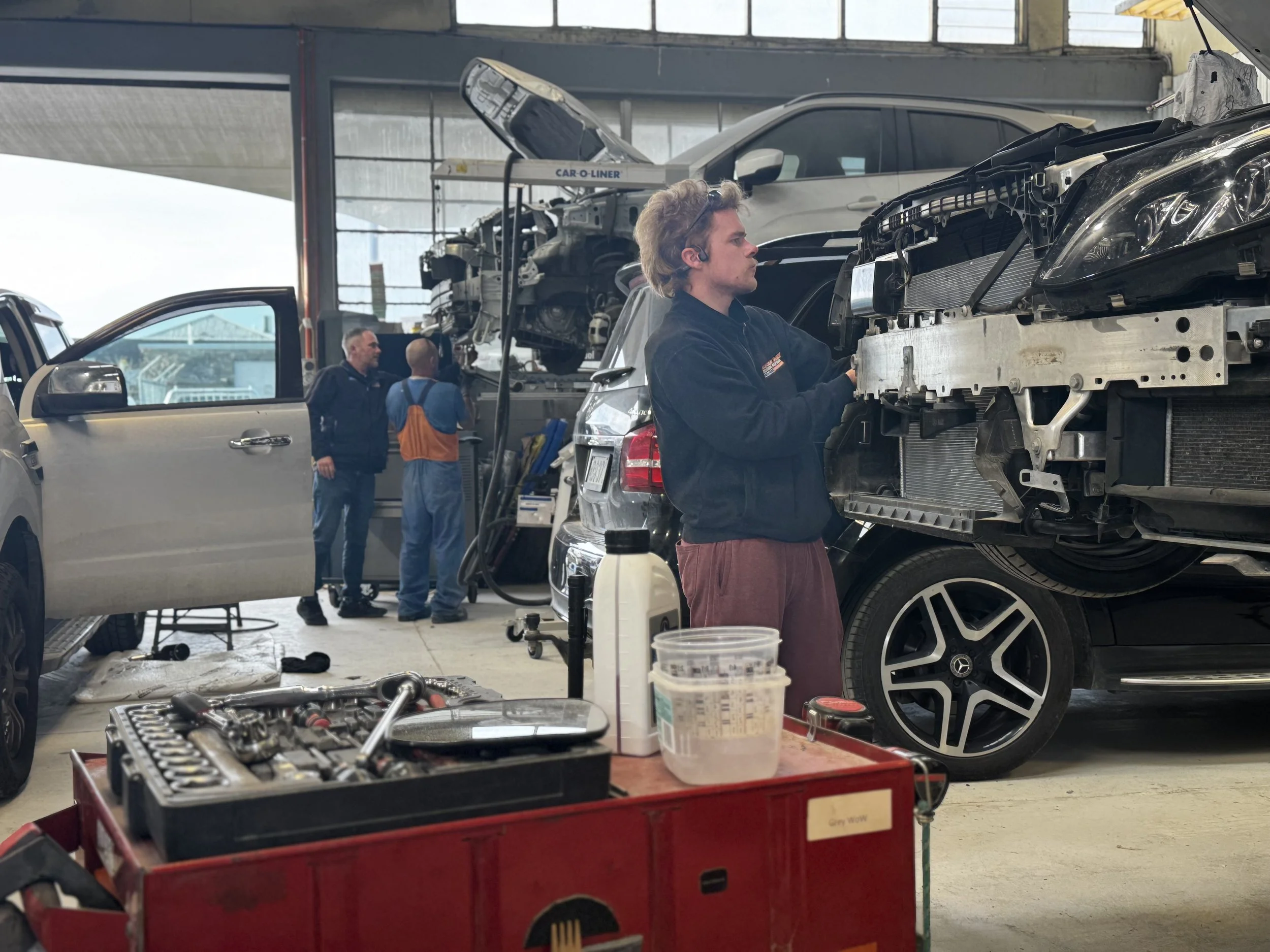 Mechanic working on a black car with its front bumper removed inside an auto repair shop. Two other cars are elevated on lifts in the background, and work tools and parts are on a workbench and in a tool box in the foreground.