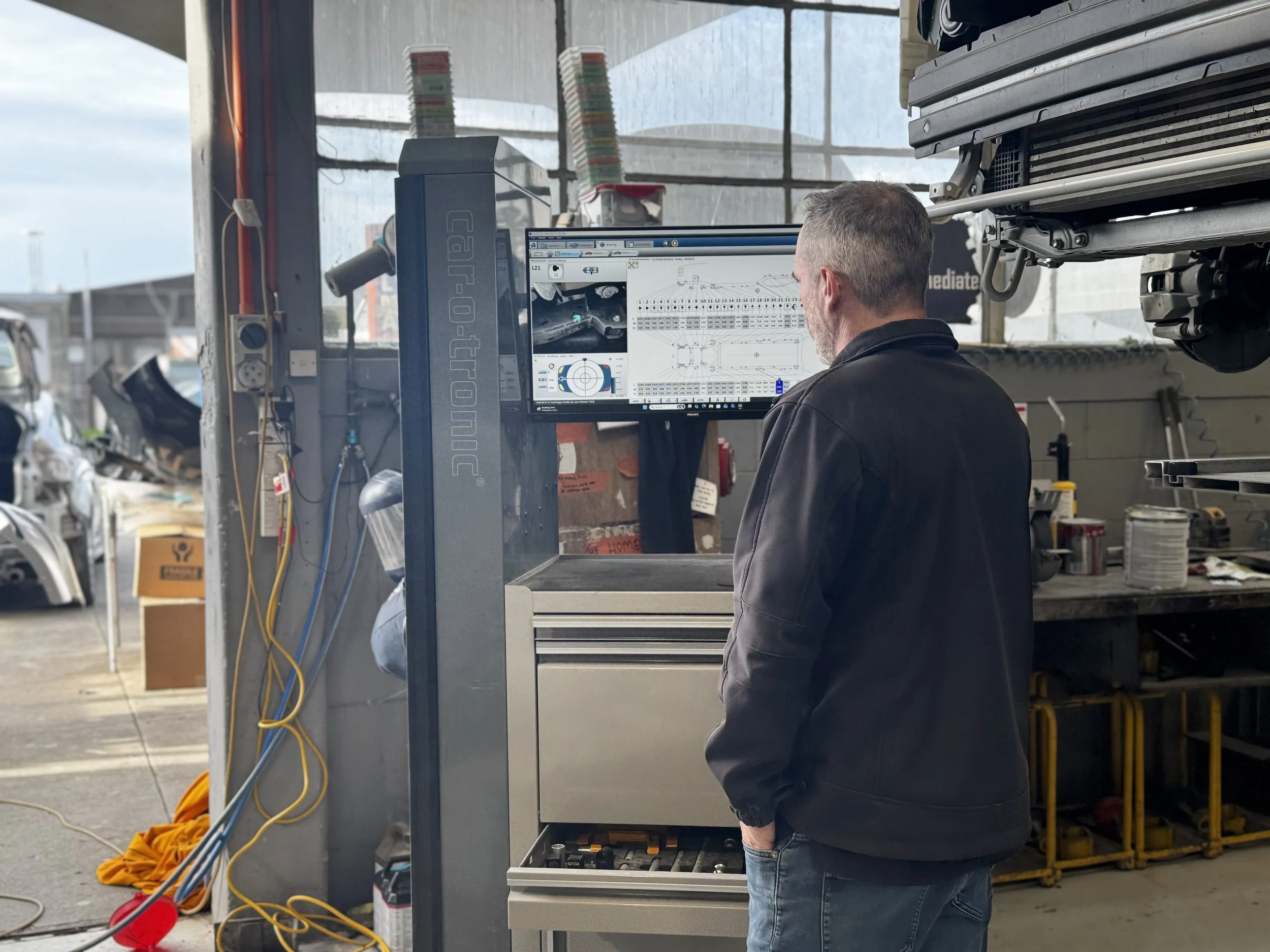 A man working on a computer inside a garage or workshop, with shelves, tools, and equipment around him, looking at a screen displaying technical diagrams and information.