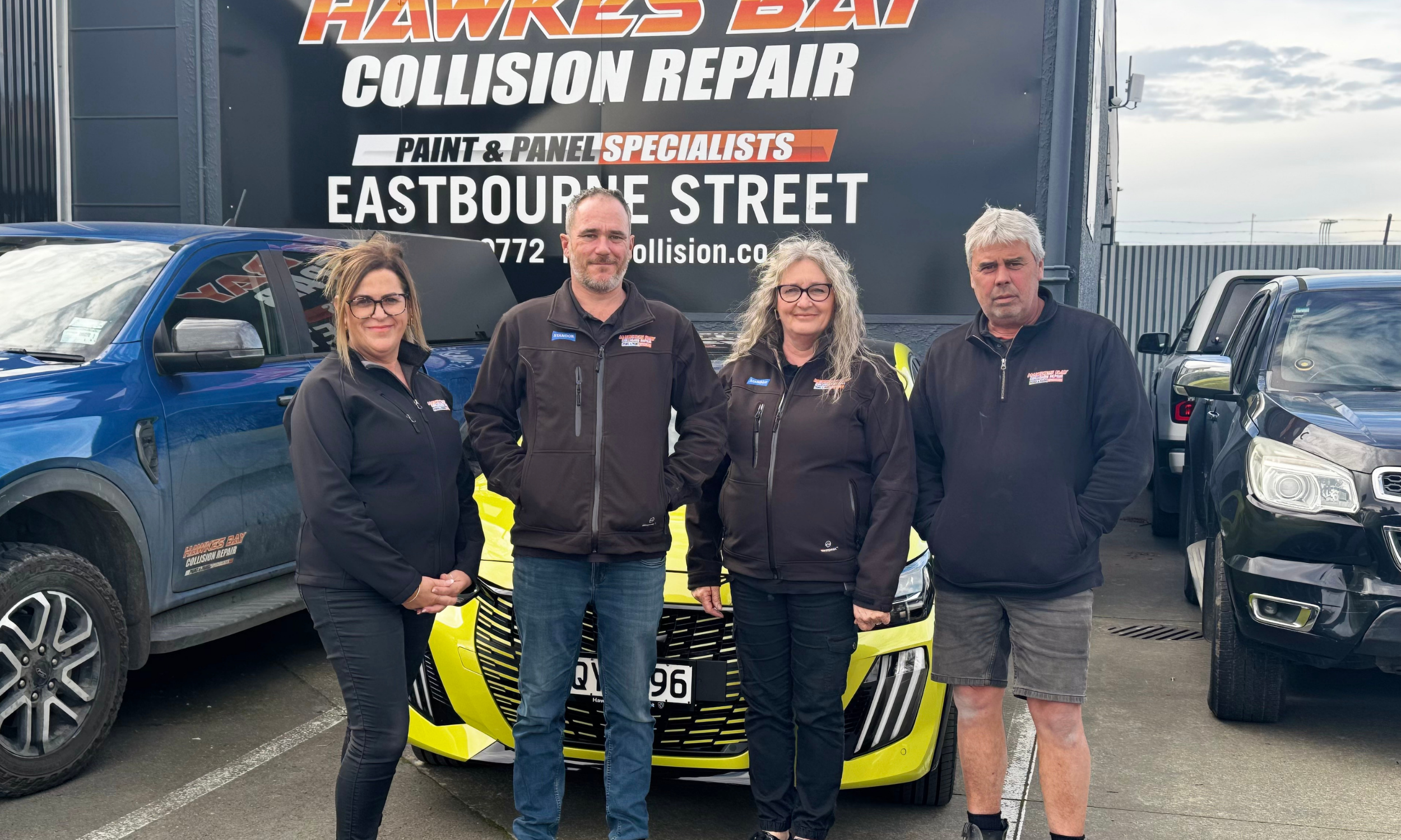 Four people standing in front of a yellow sports car and a building with a sign for Hawkes Bay Collision Repair. The group includes two women and two men, all wearing black jackets with the company's logo.