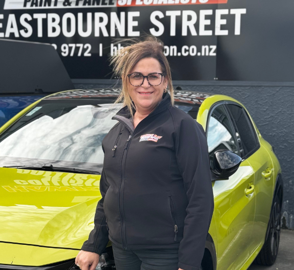 Women in black jacket standing next to a lime green car with black wheels, in front of a black wall with white and red signage.
