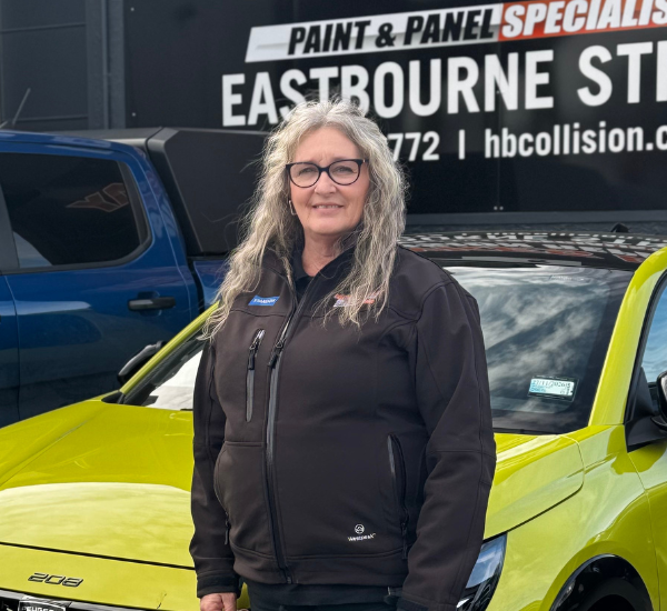 Woman with glasses and long gray hair standing in front of a yellow car and a building with a sign for Eastbourne Street paint and panel specialists.